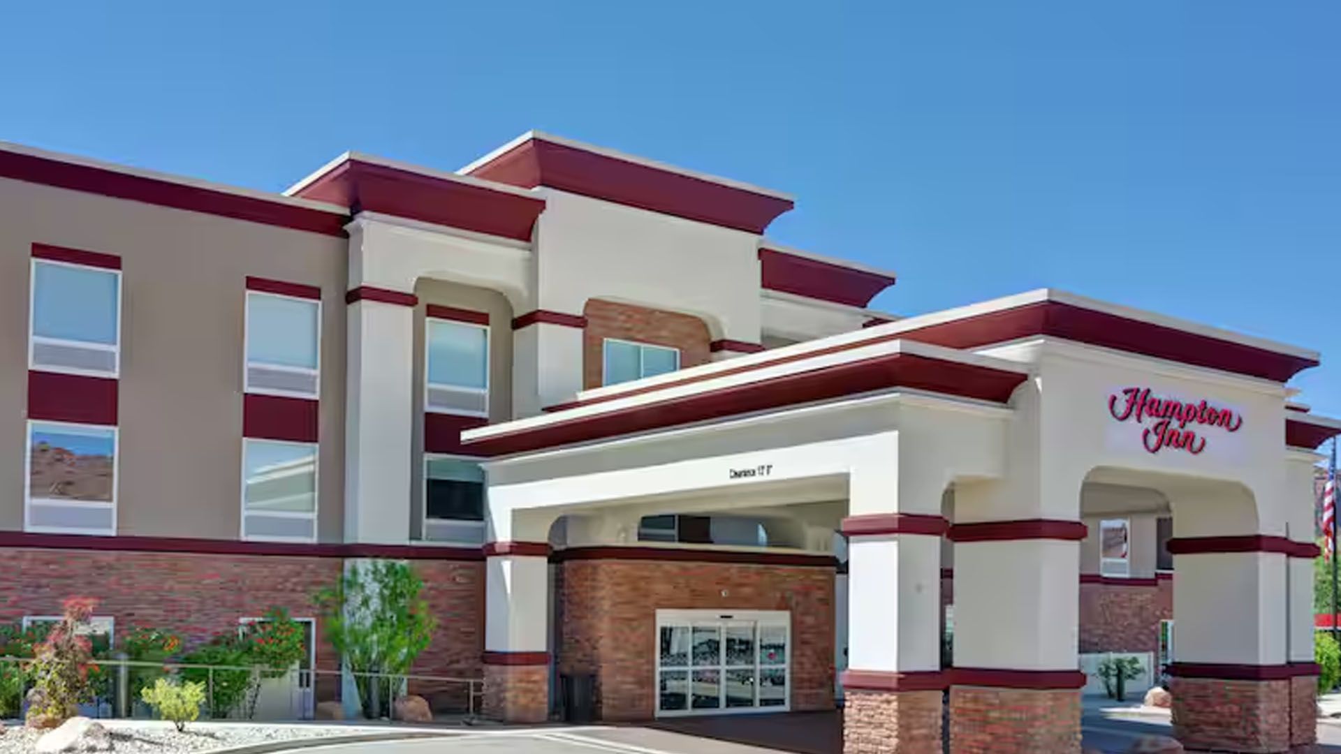 Hampton Inn hotel entrance with red trim and brick facade under a clear blue sky.