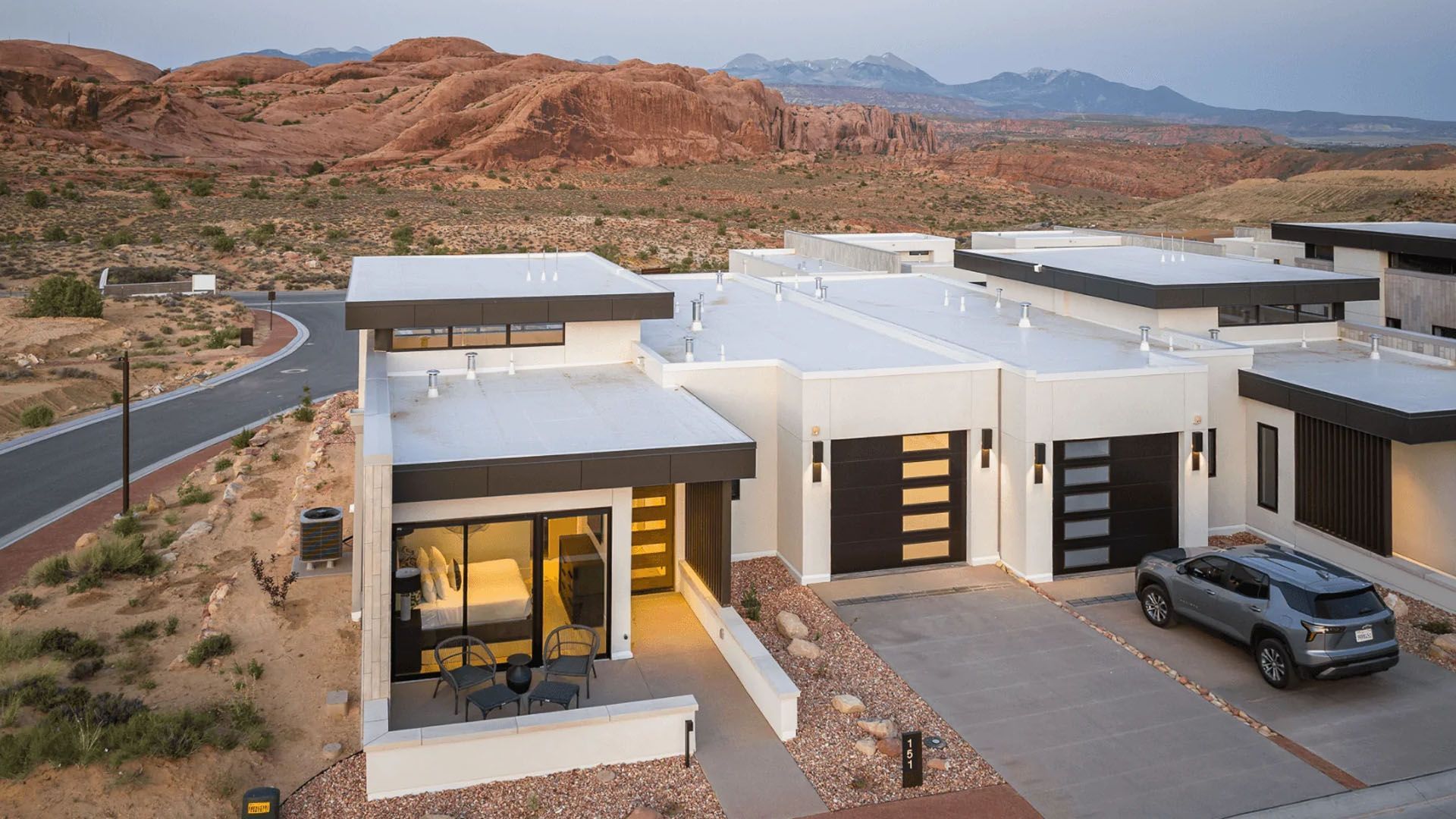 Modern white house with black accents in a desert setting, a car in the driveway, and mountains in the background.