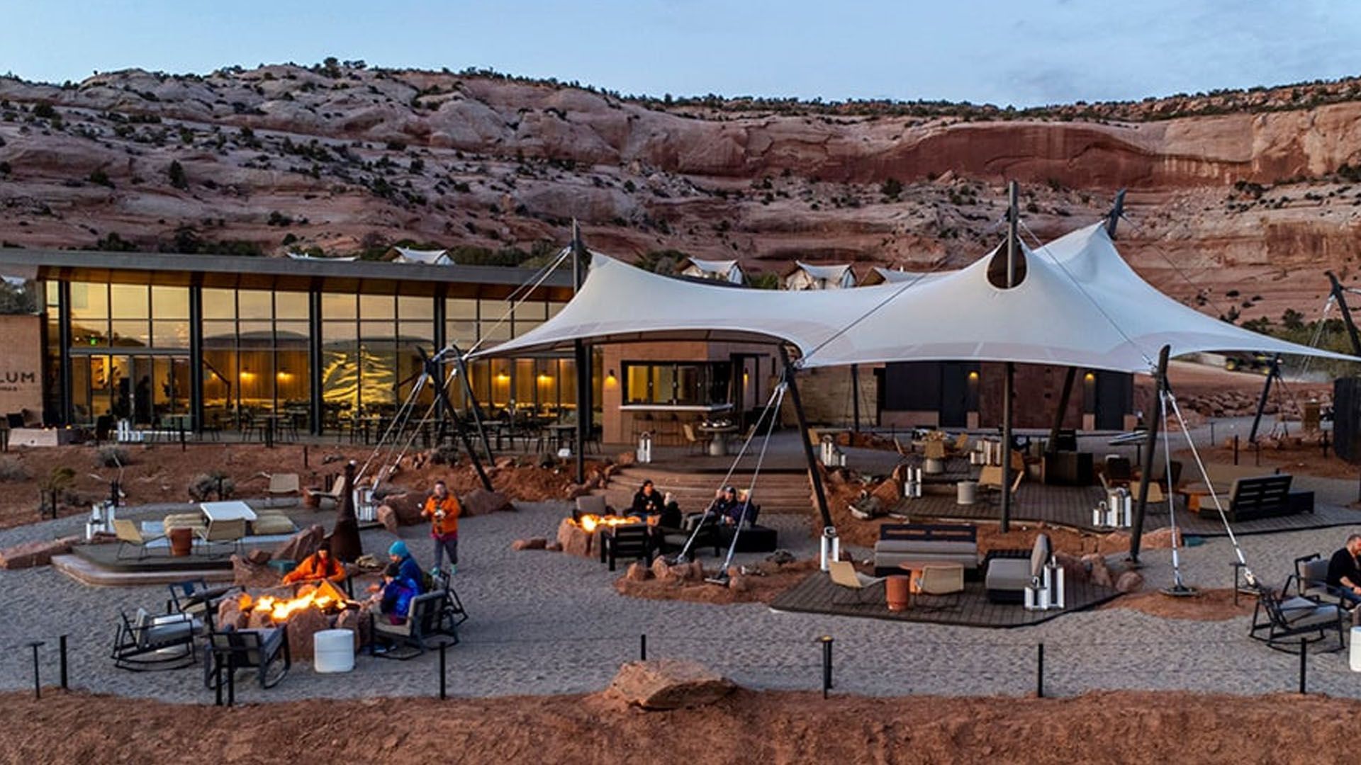 Outdoor desert lodge with a tent, fire pit, and people seated under a canopy, at dusk.