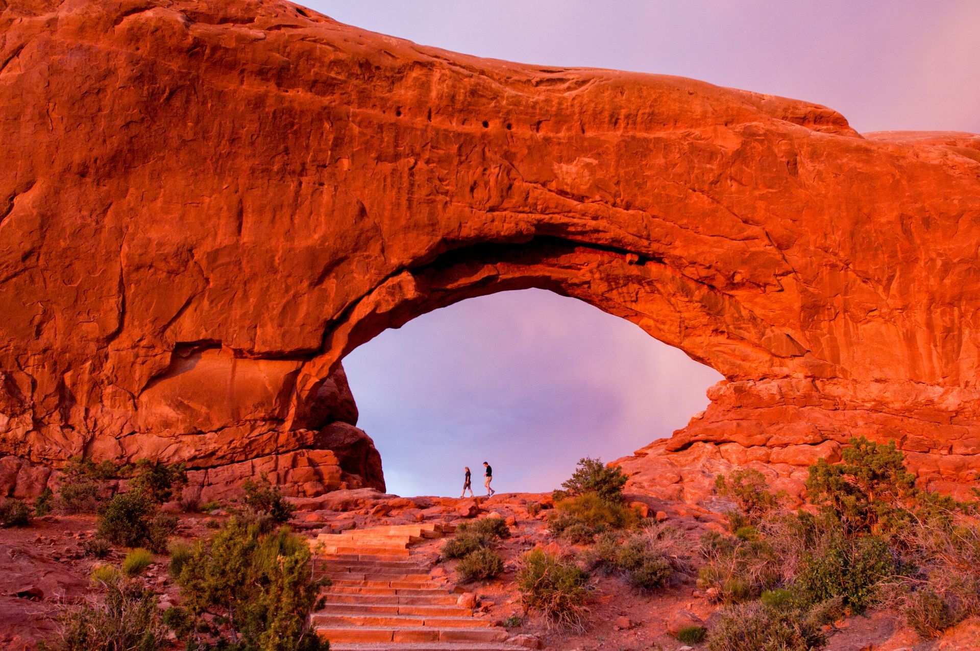 Red rock arch with two people silhouetted in the opening; sunset colors.