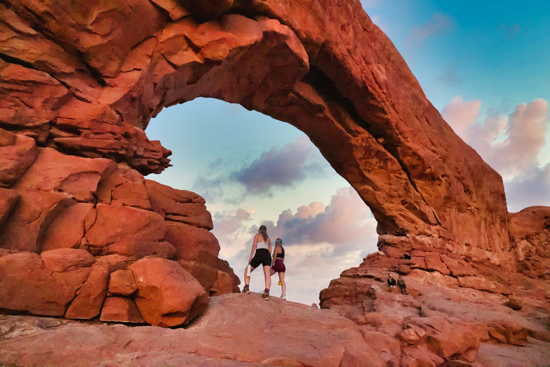 Two people stand under North Window Arch against a blue sky with clouds.