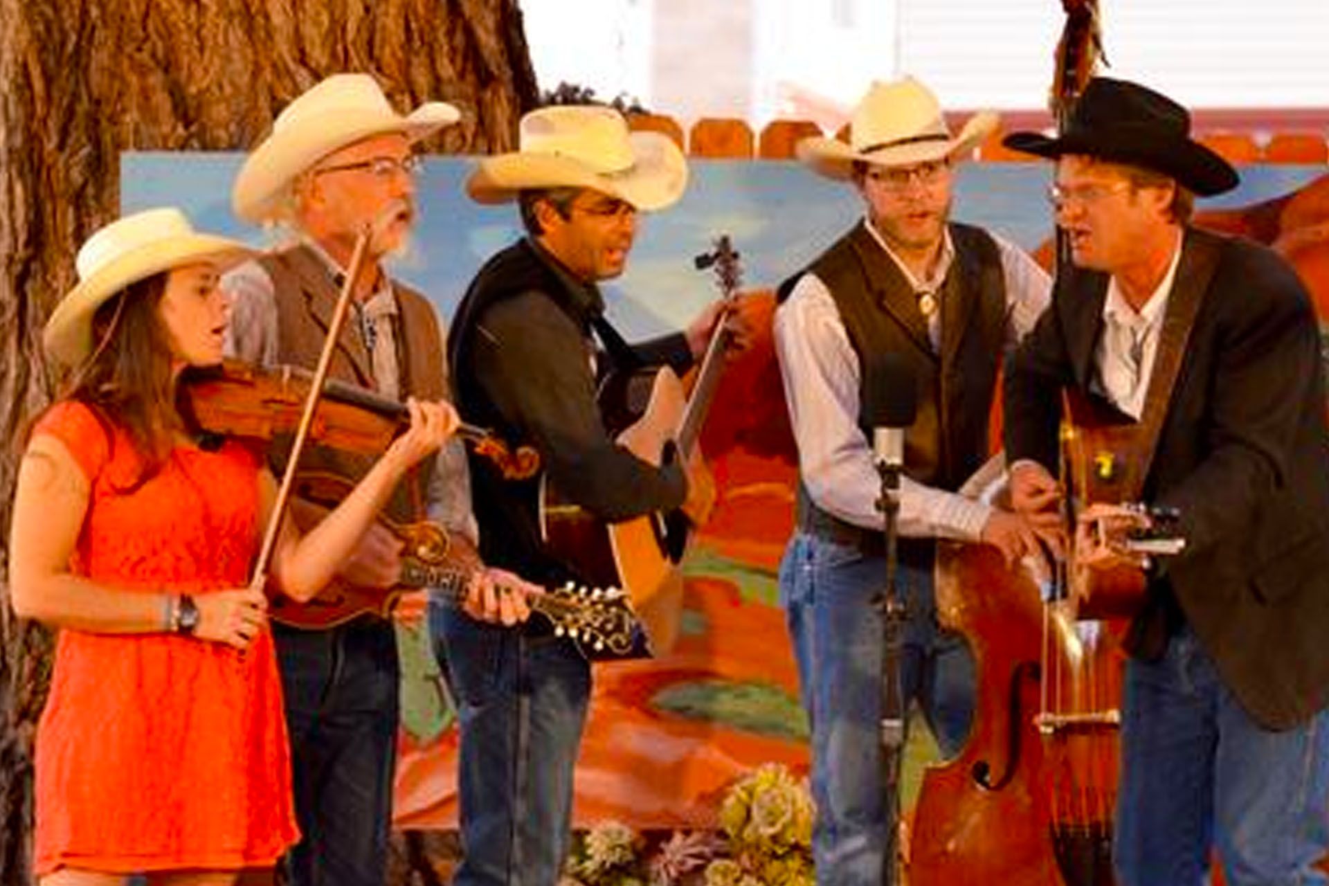 A band of six musicians in cowboy hats perform outdoors; a woman plays the violin, others play guitar and upright bass.