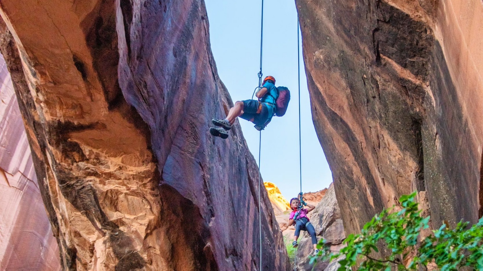 Two people rappelling down a narrow canyon; red rock walls and blue sky.