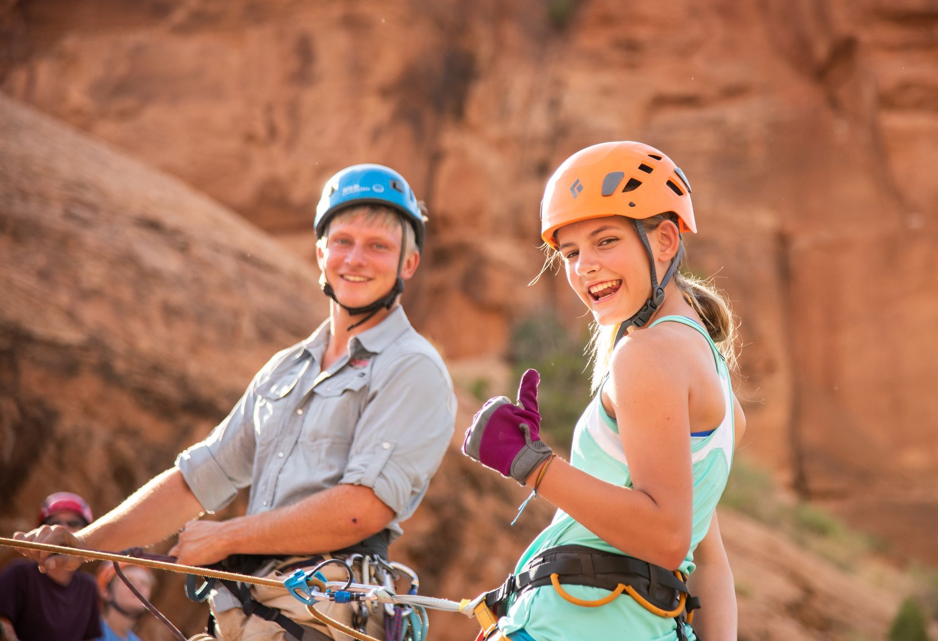 Climbing guide and girl in climbing gear, smiling, thumbs up, in front of a red rock cliff.