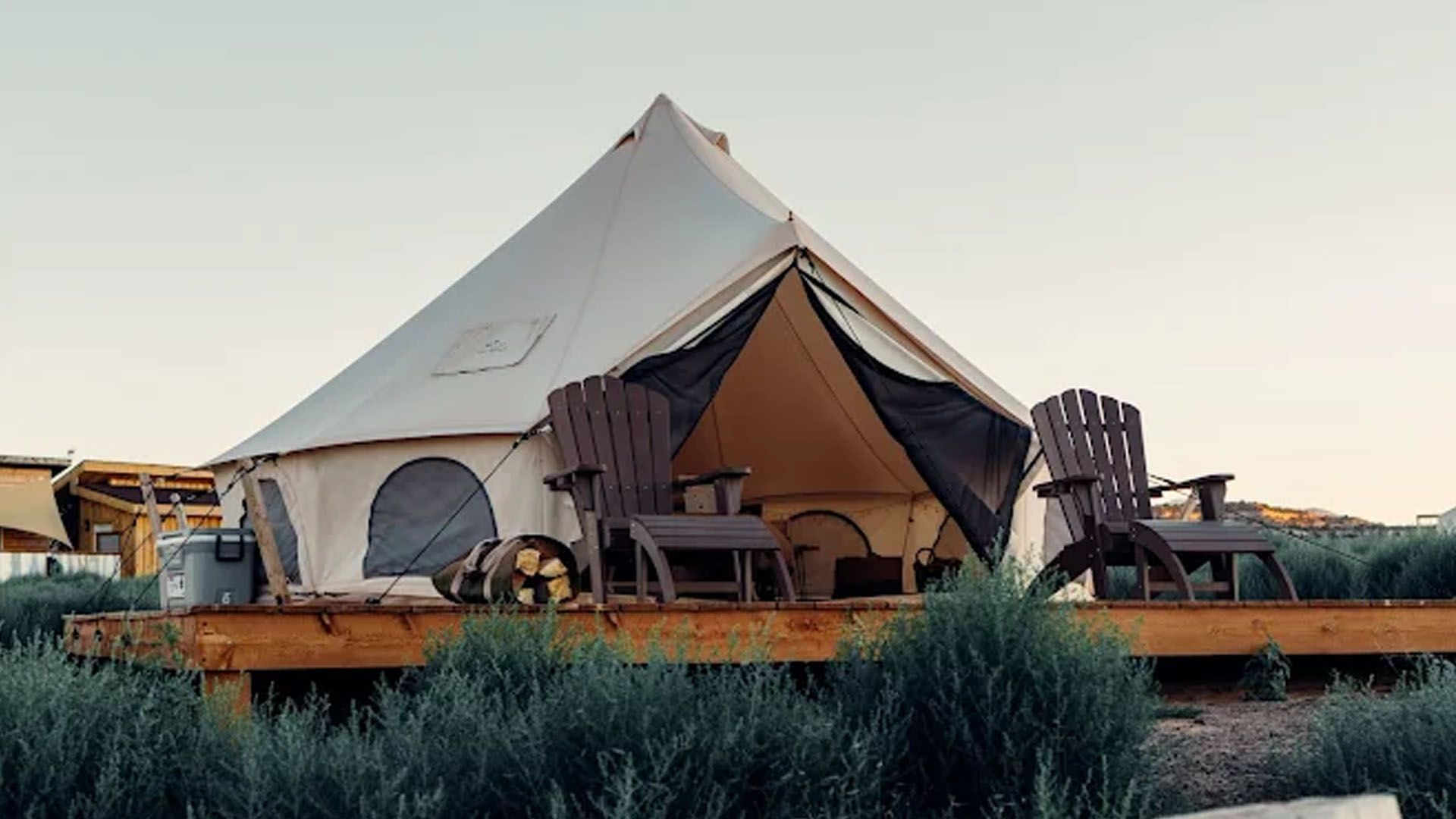 Canvas tent with two wooden chairs on a wooden deck surrounded by greenery, in an outdoor setting.