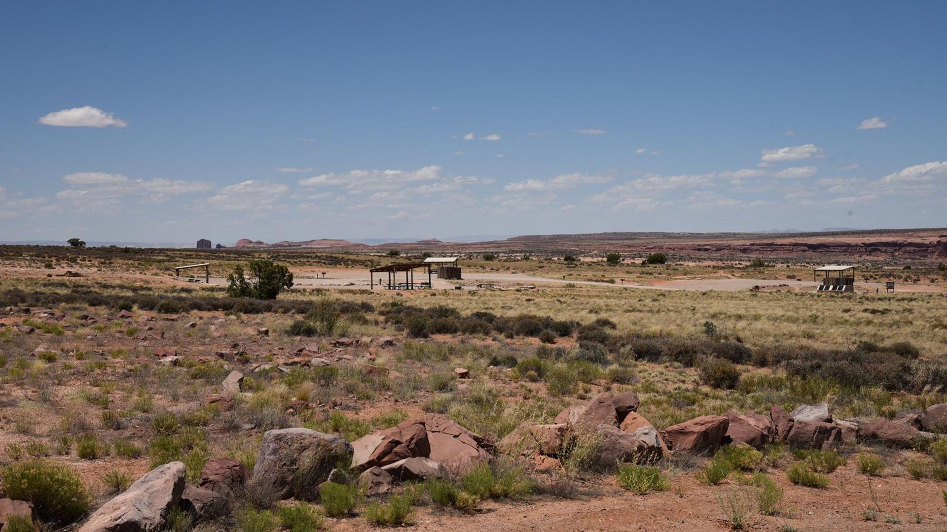 Desert landscape with campsite shelters under a blue sky. Red rock and sparse vegetation.