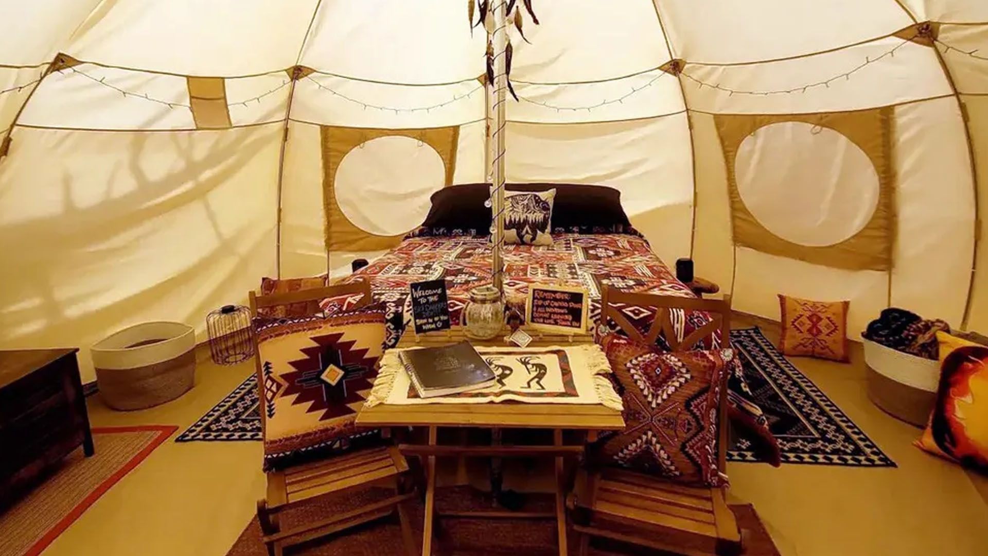 Inside a glamping tent: Bed with decorative pillows, small table with chairs, rugs, and baskets.