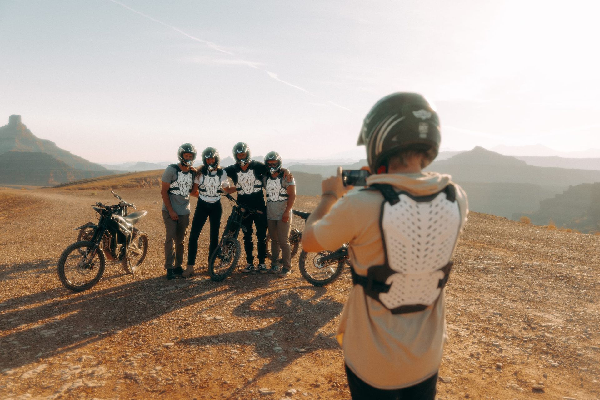 Person taking photo of four people with dirt bikes on a desert hillside. They all wear helmets and body armor.