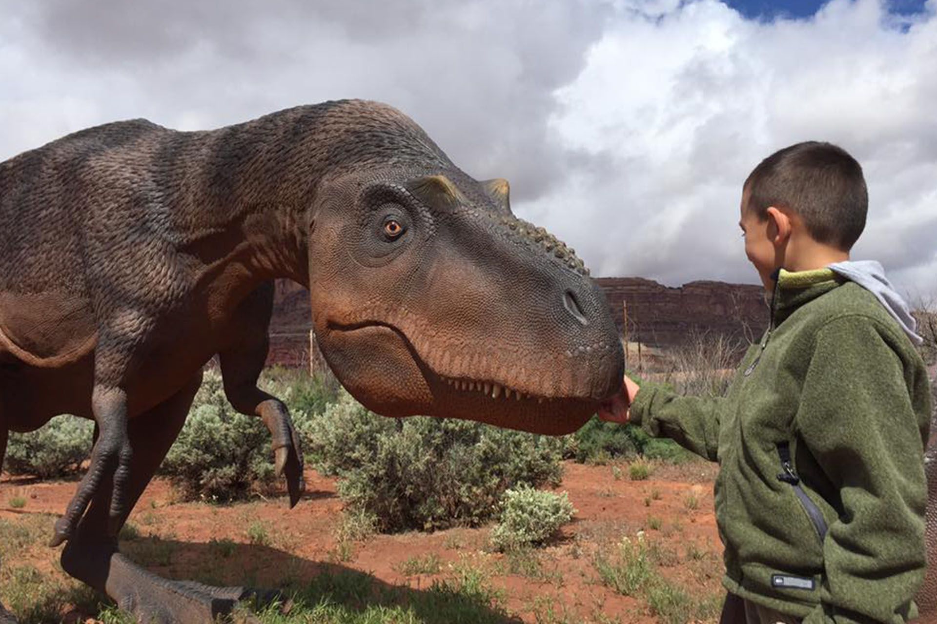 Young boy appears to be petting a life-size T-Rex in the outdoors