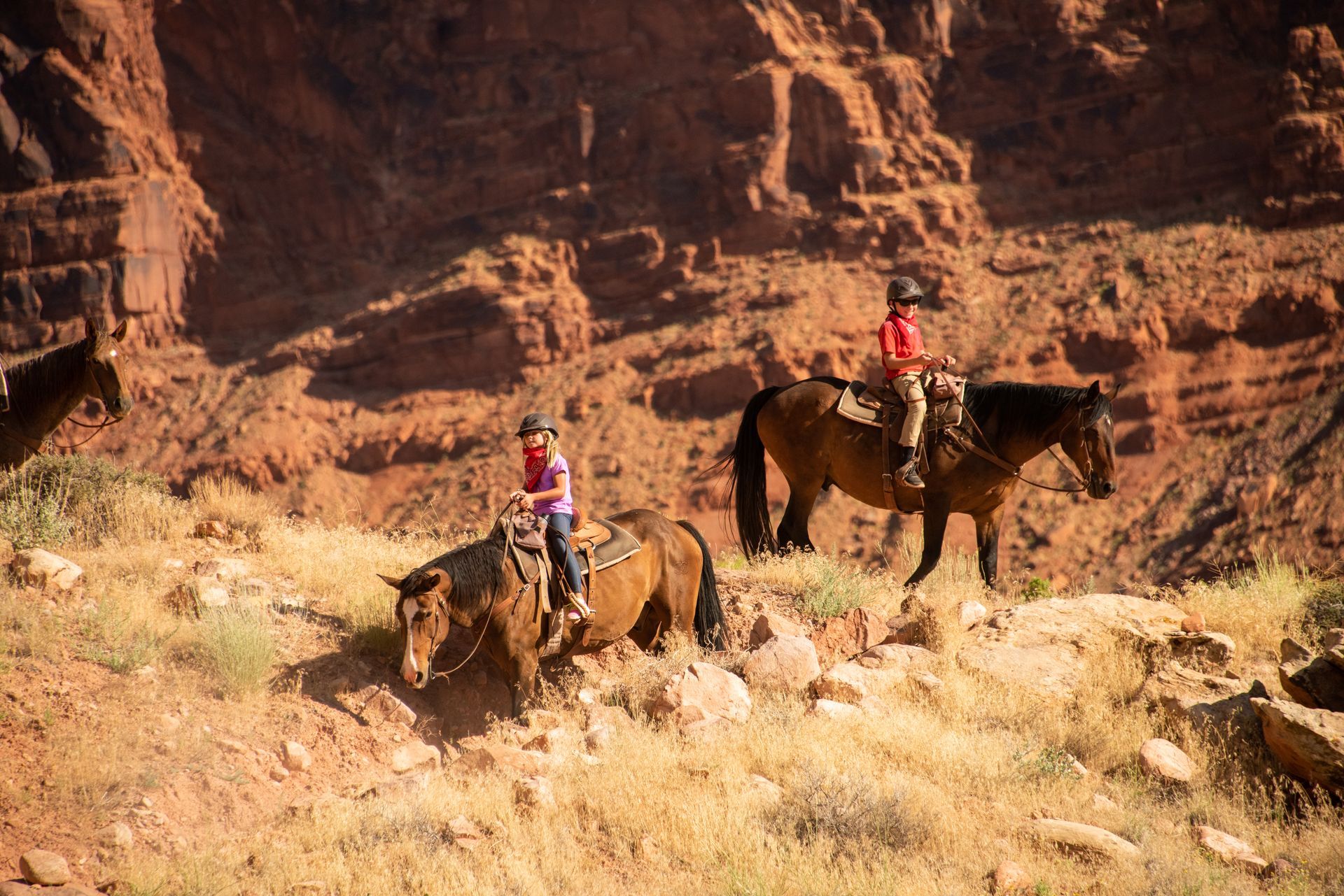 Two children horseback riding on a trail in a red rock canyon.