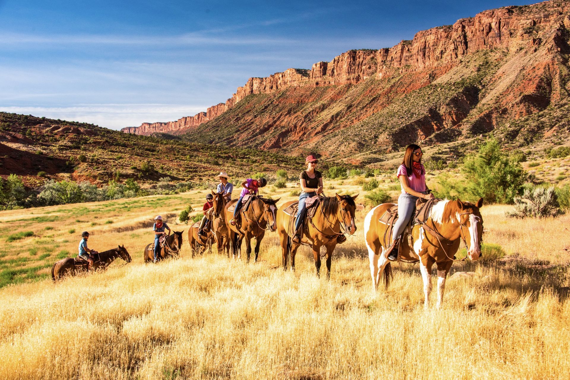 People horseback riding through a field with red rock formations in the background.
