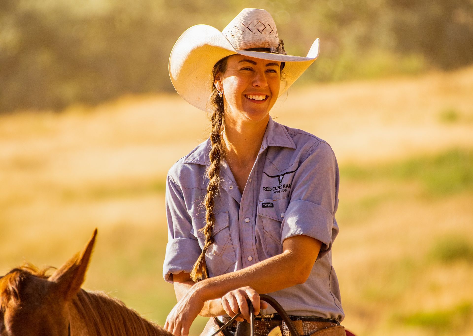 Wrangler smiles while riding a horse in a sunny field, wearing a cowboy hat and blue shirt.