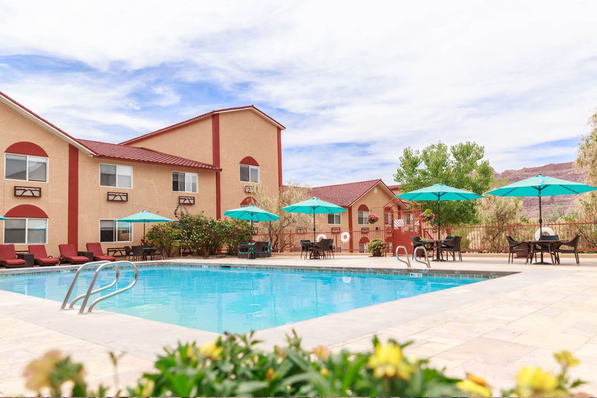 Swimming pool with turquoise umbrellas and lounge chairs in front of a beige hotel; sunny day.