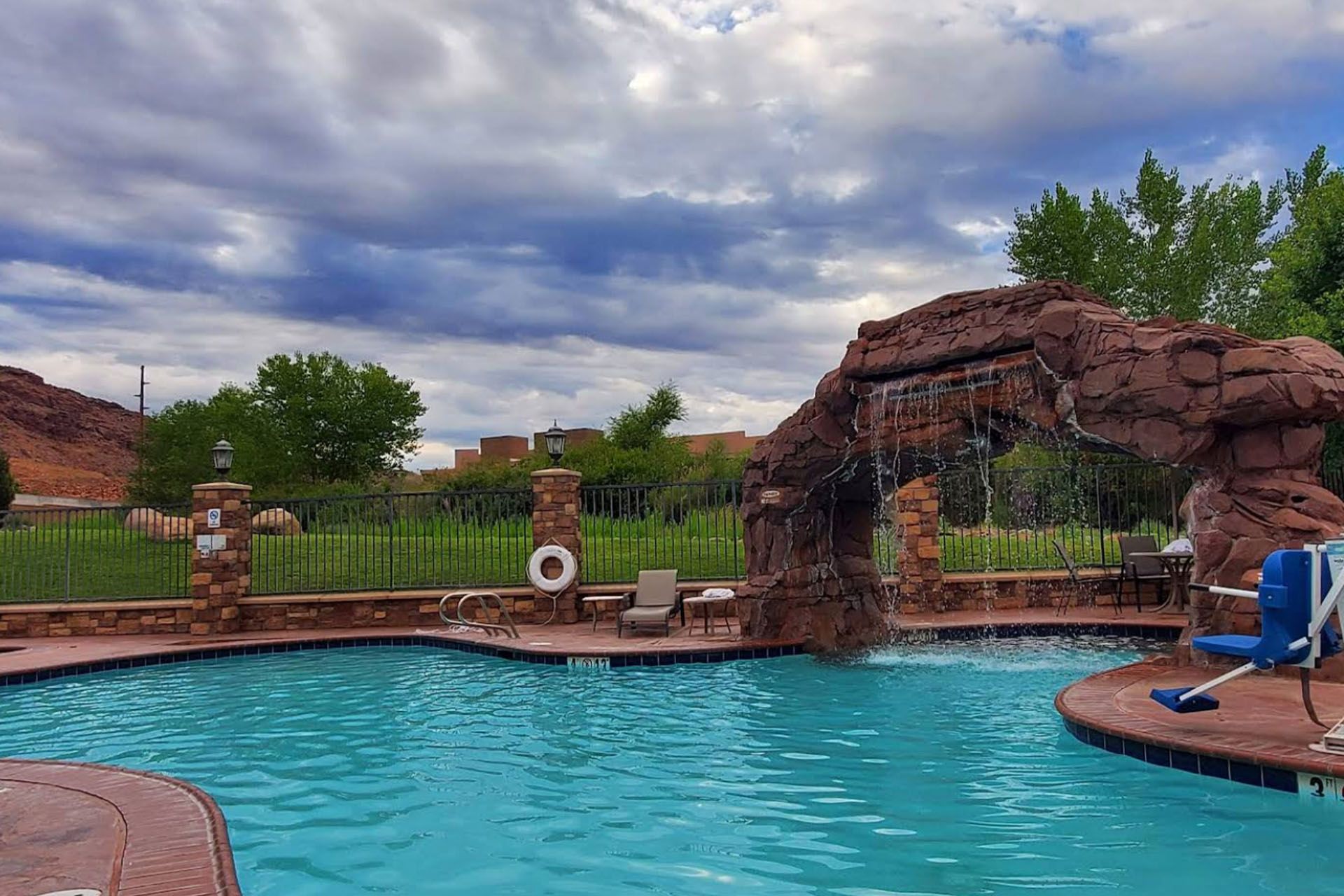 Pool with a rock waterfall feature, surrounded by a fence and trees. Cloudy sky.