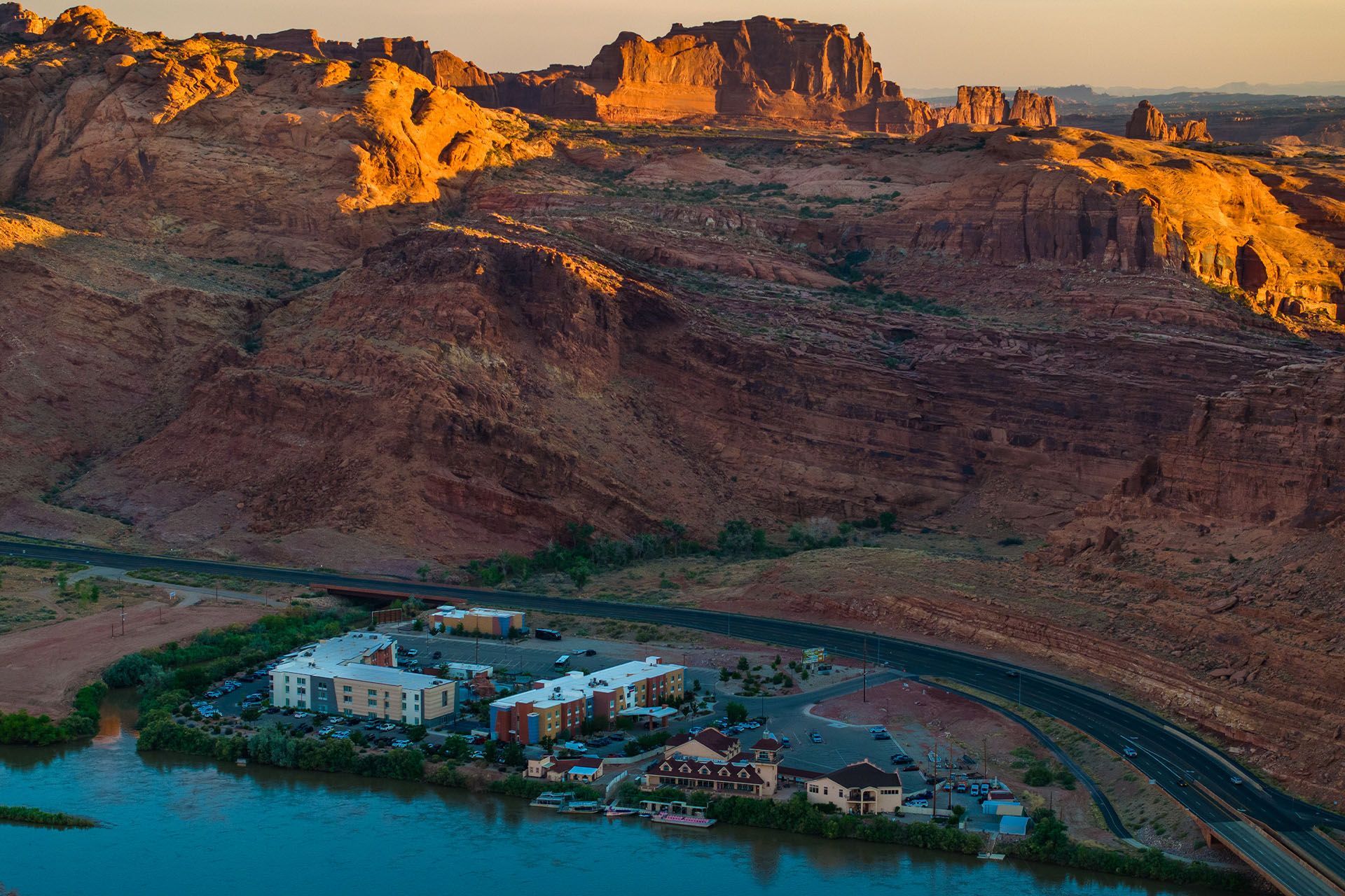 Hotel complex nestled by river, contrasting with red rock mountains in the background under warm sunlight.