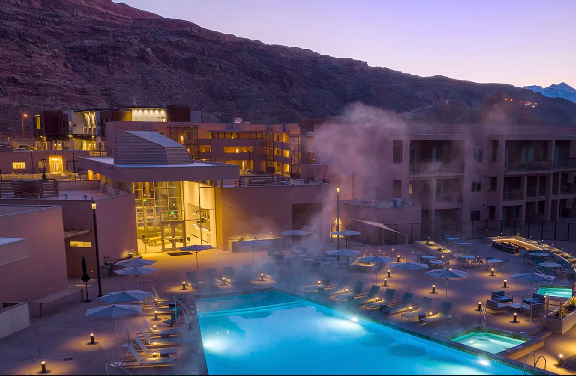 Luxury resort with lit pools and steam, nestled in a mountain valley at dusk.