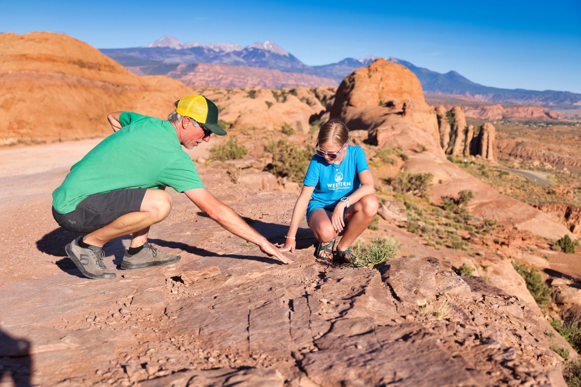 Man and girl examining rock surface outdoors with mountains in background.