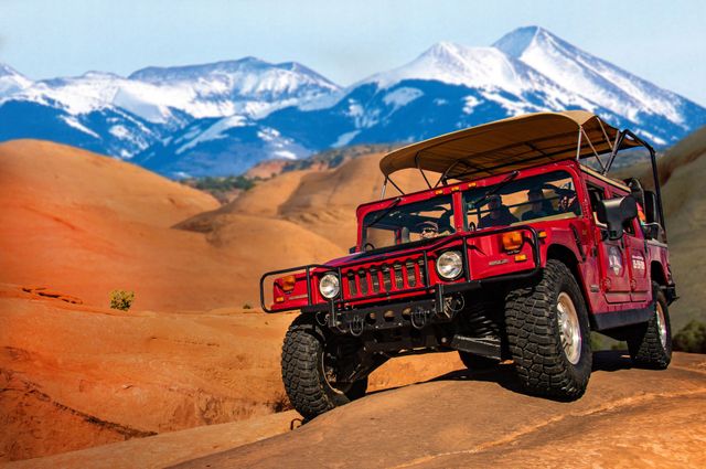 A Hummer vehicle drives across a sandstone fin with the La Sal Mountains in the background.
