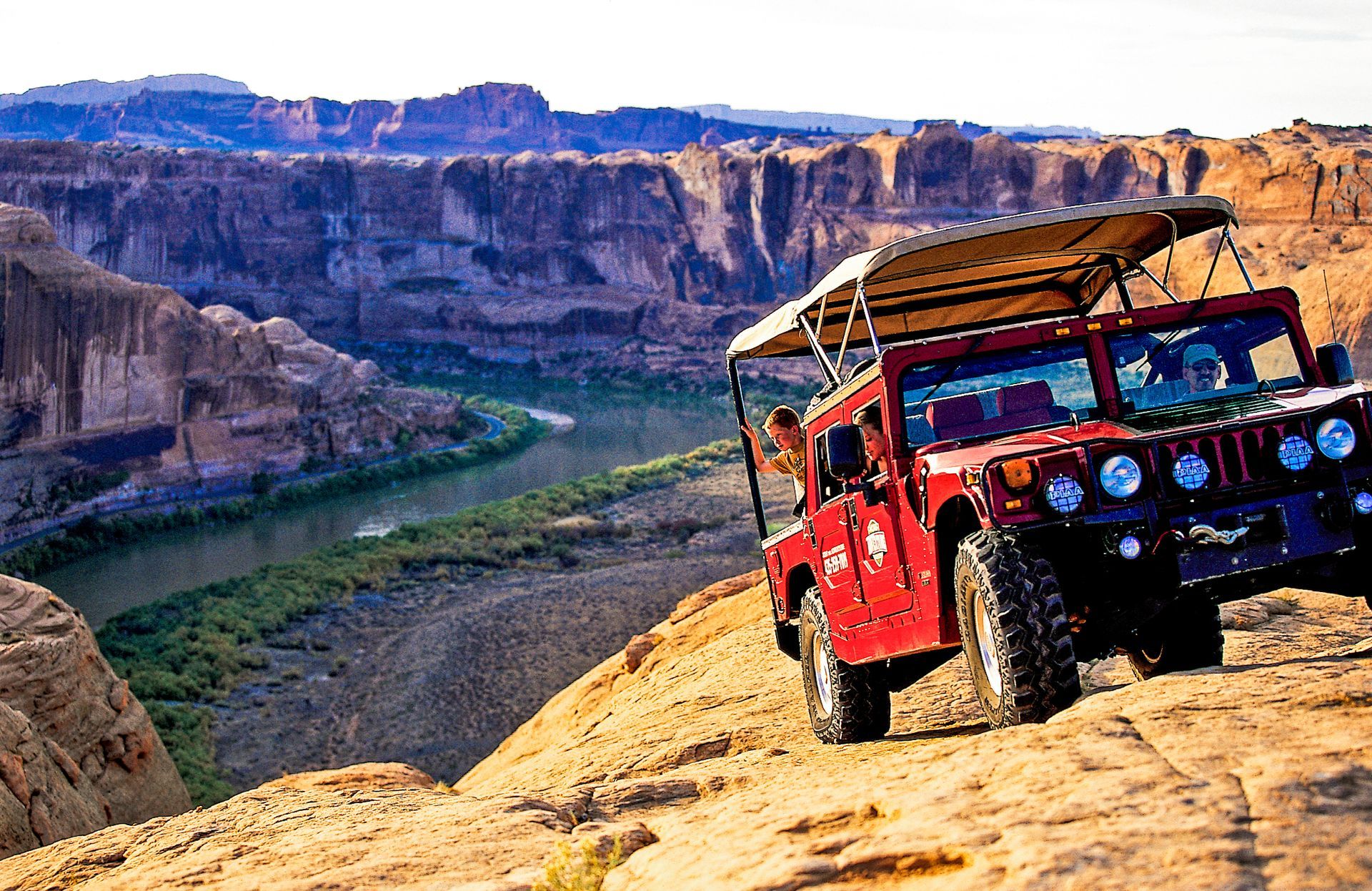 An H1 Hummer drives on a rocky cliff, with a river and canyon in the background.