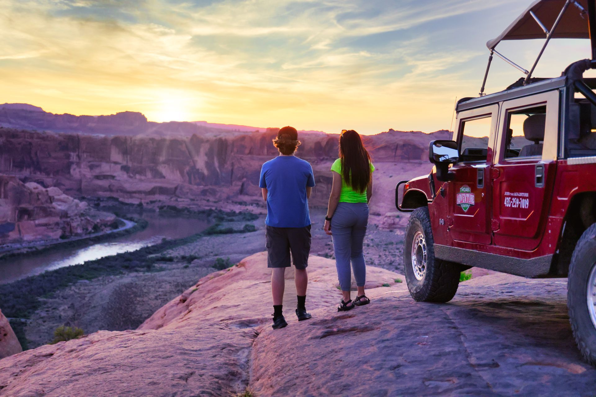 A man and a woman are standing on top of a rock next to a jeep.