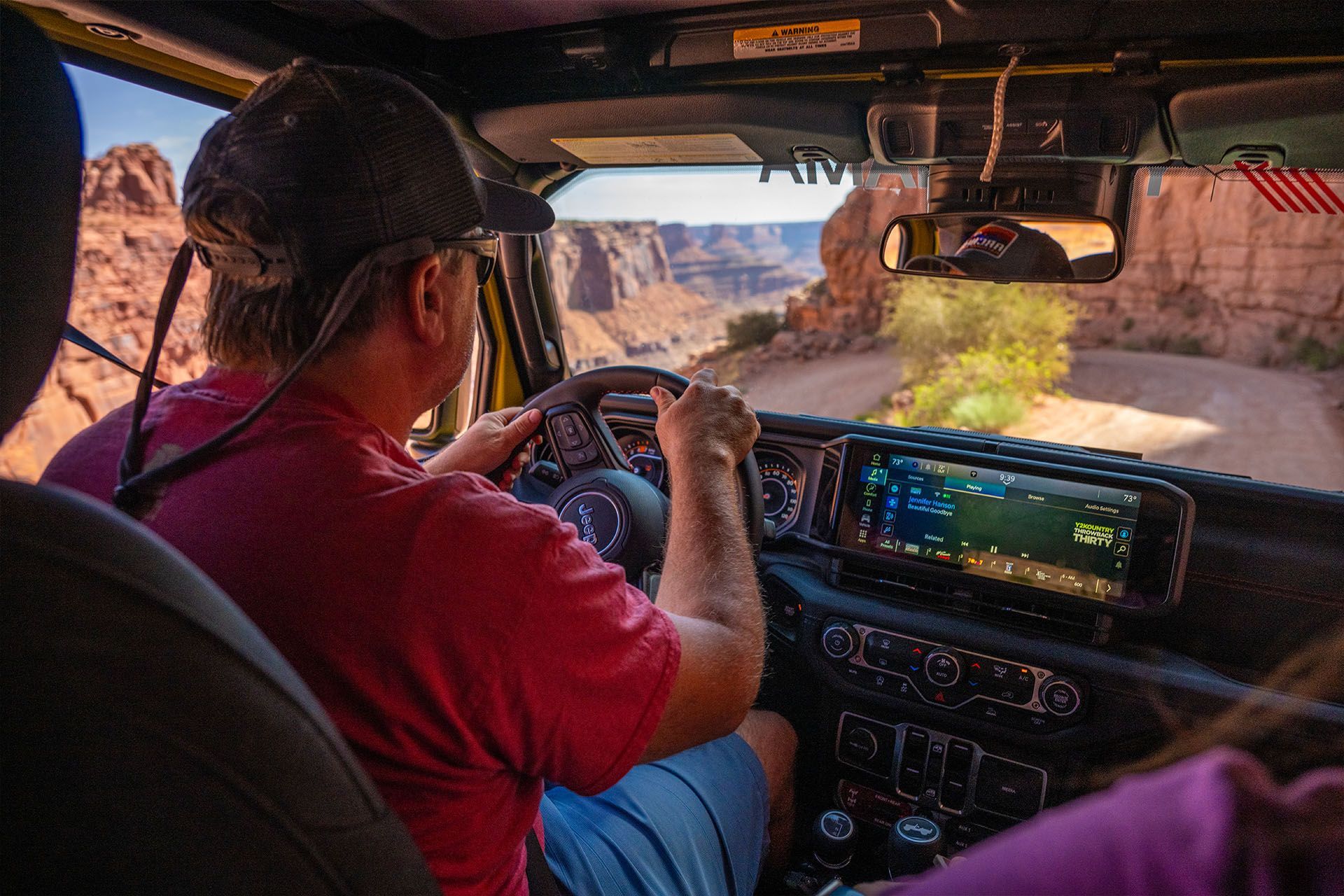 Man driving a Jeep on a dirt road through a canyon, navigation screen on, bright sunny day.