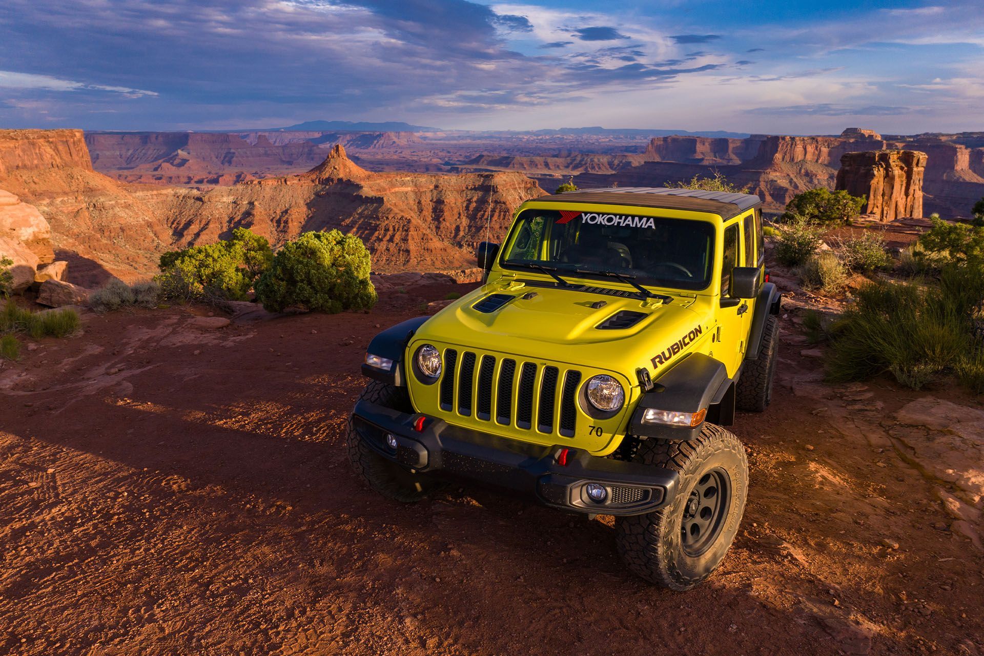 Yellow Jeep parked on a red-dirt cliff, overlooking a canyon. Cloudy sky in background.