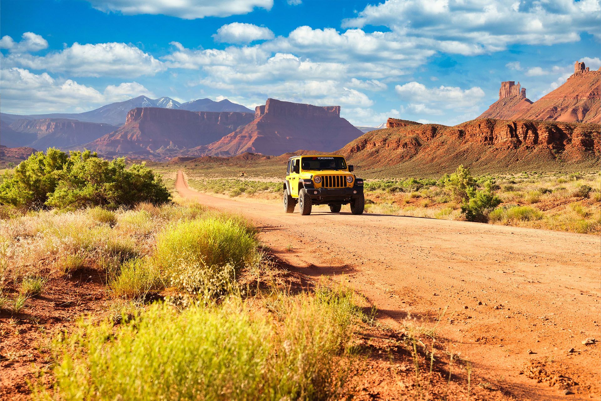 Yellow Jeep drives on a dirt road in a desert landscape with red rock formations and blue sky.