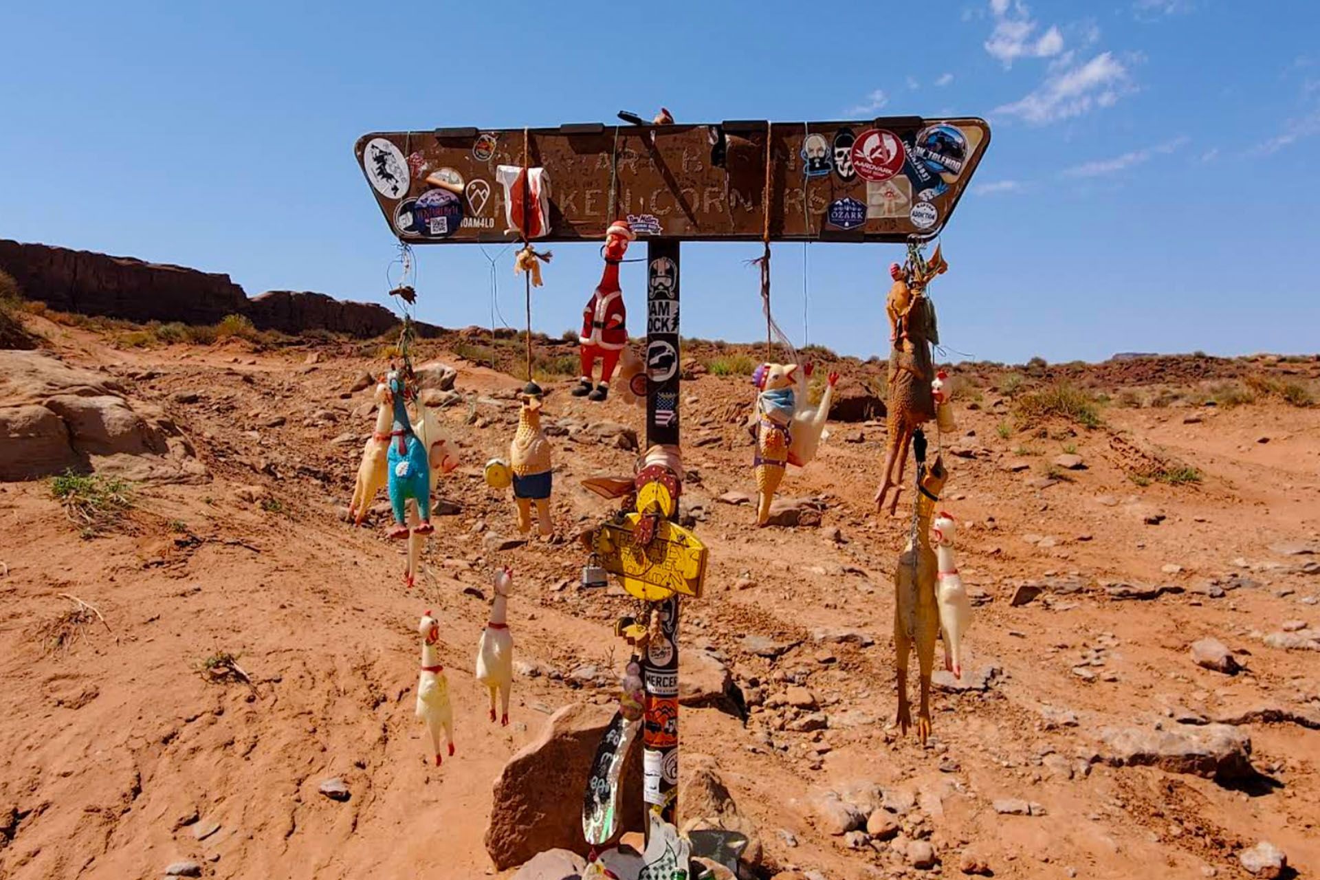An off-road trail sign for chicken corners is covered in stickers and has rubber chickens hanging from it.