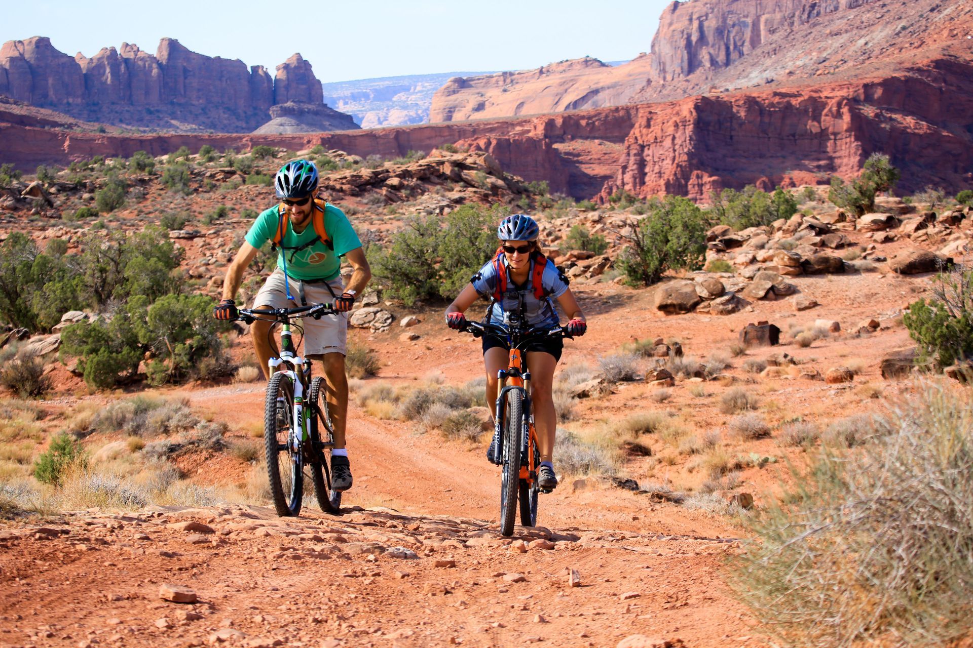Two people mountain biking on a red dirt trail in a desert landscape with red rock formations.