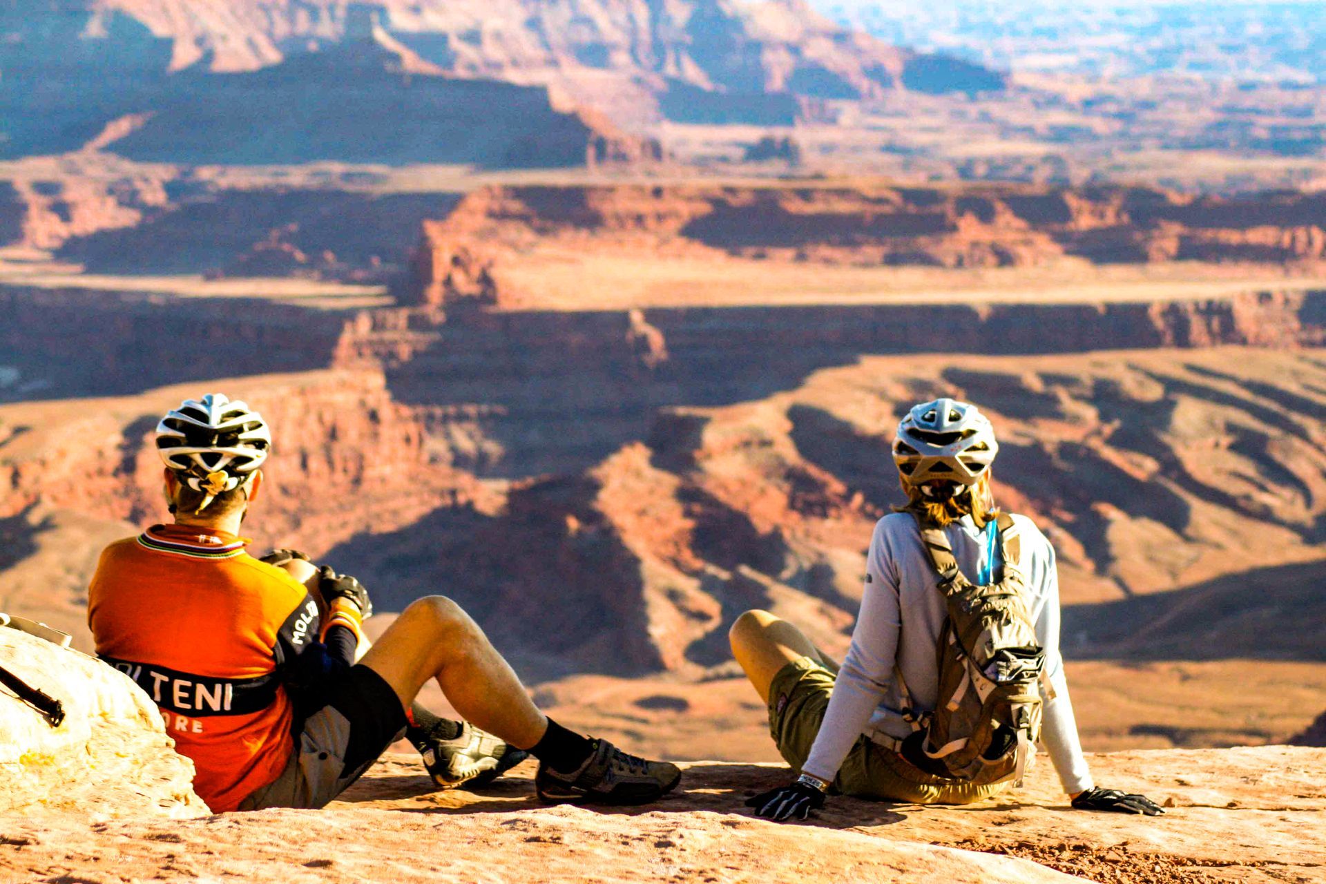 Two people sit with bike helmets on as they look over vast canyons from an overlook.