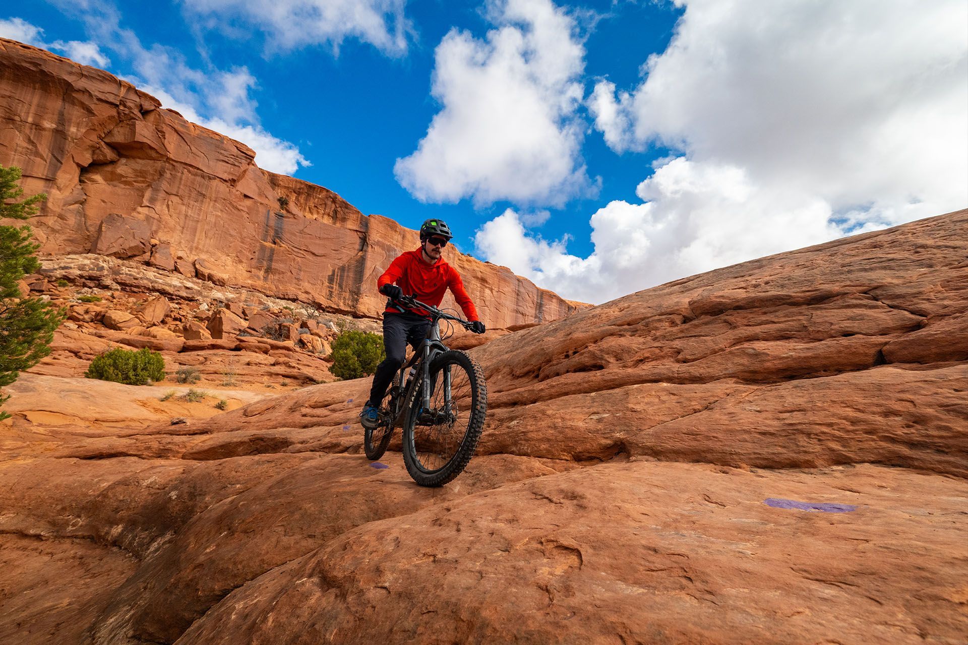 A man in a red shirt and black pants rides a mountain bike across slickrock terrain.