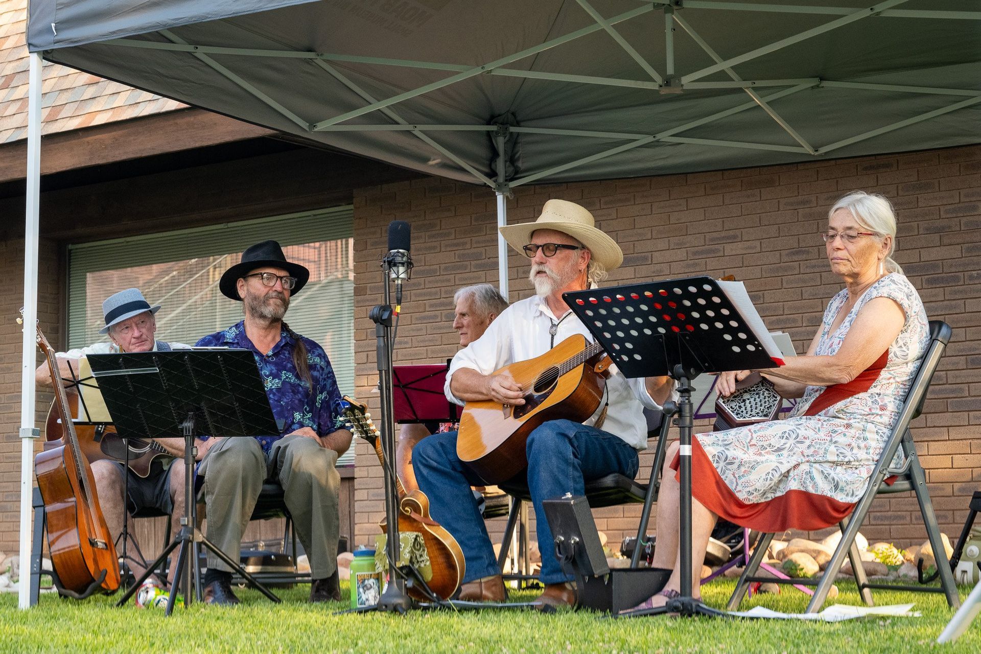 A band of five people plays music outdoors under a canopy. They're sitting and playing instruments.