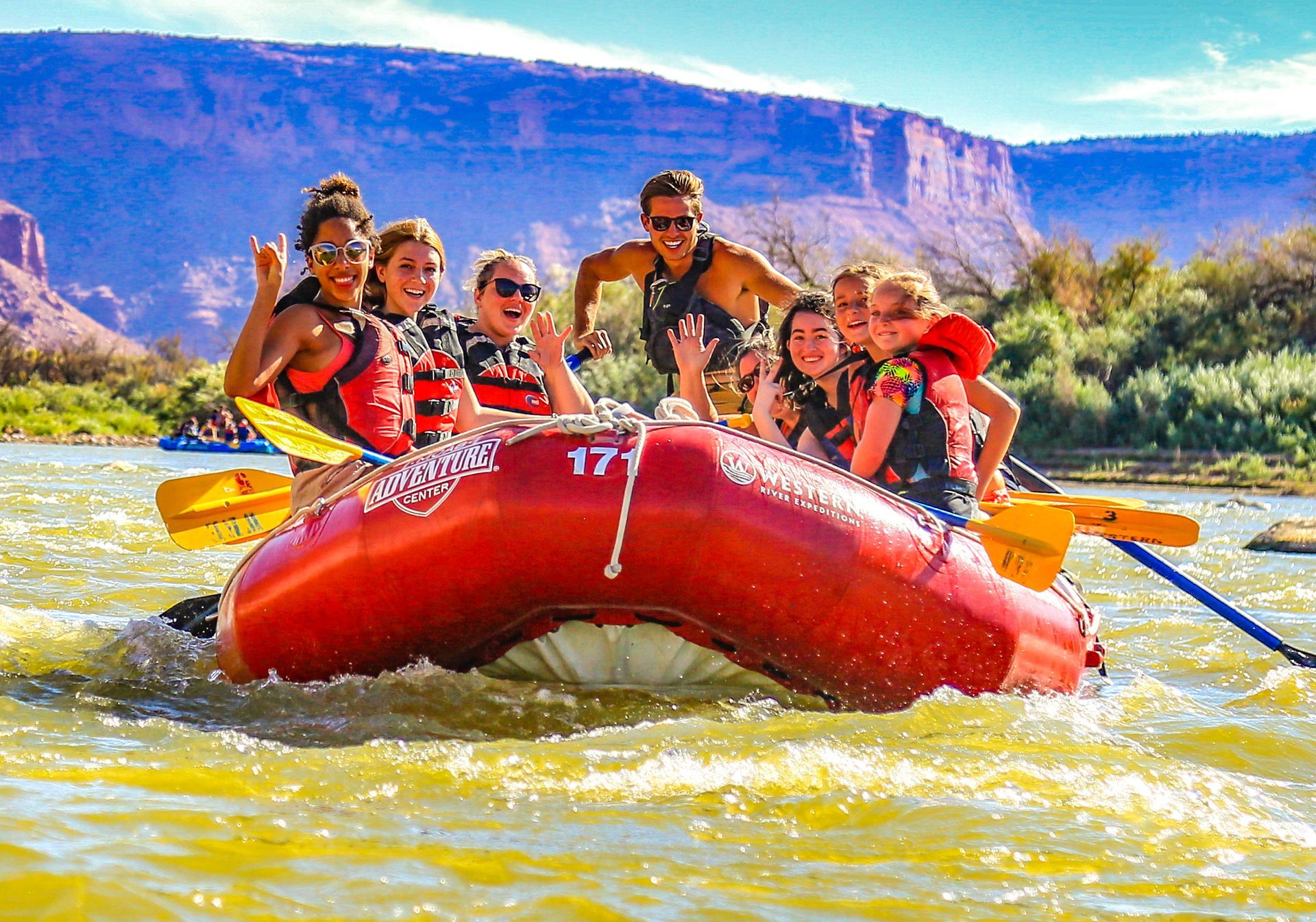 A youth group is in a raft smiling as they float downriver with cliffs in the background.