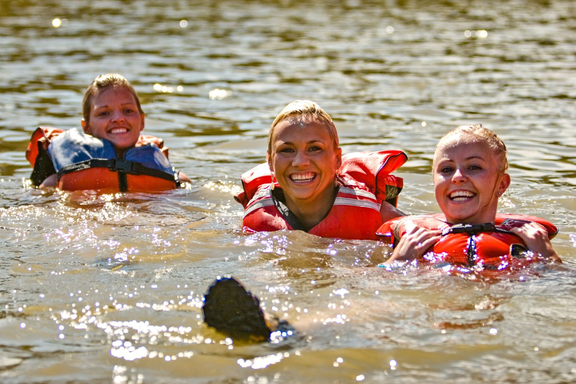 Three women in orange life vests swimming and smiling in a body of water.