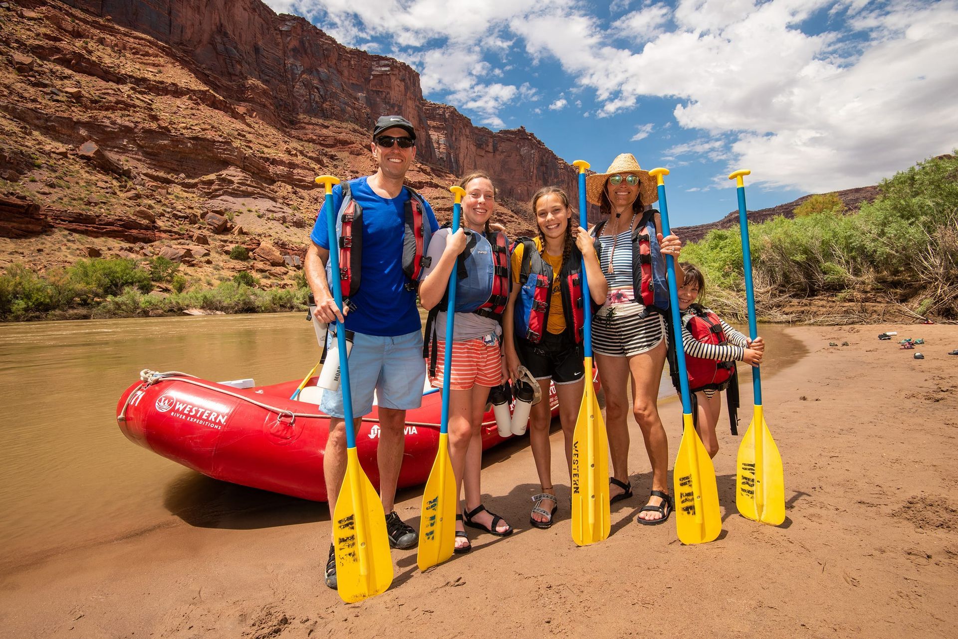 Family with paddles stands by a red raft on a sandy riverbank, with red rock canyon in background.