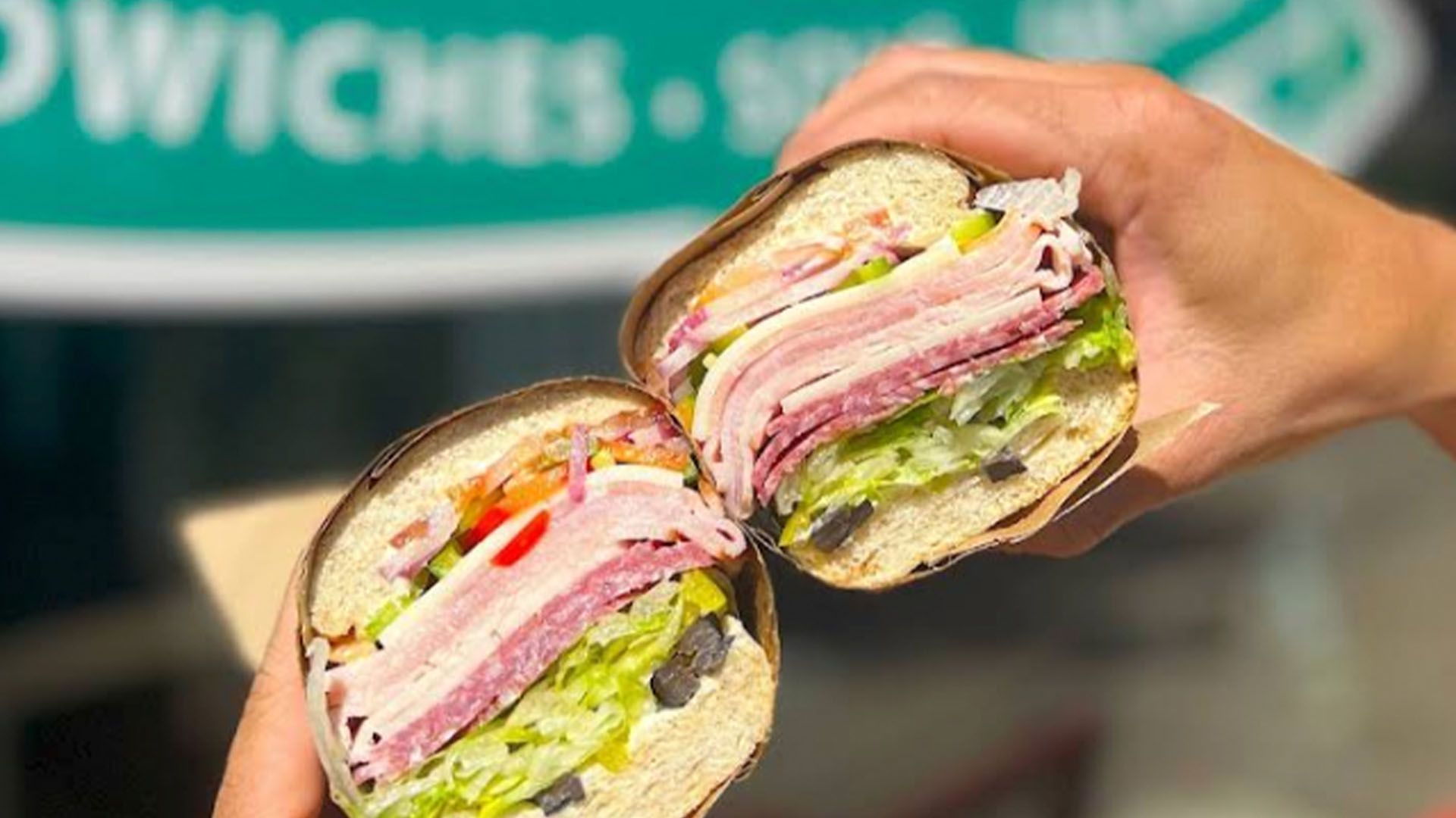 Two halves of a loaded submarine sandwich held by hands, showing meats, lettuce, and olives.