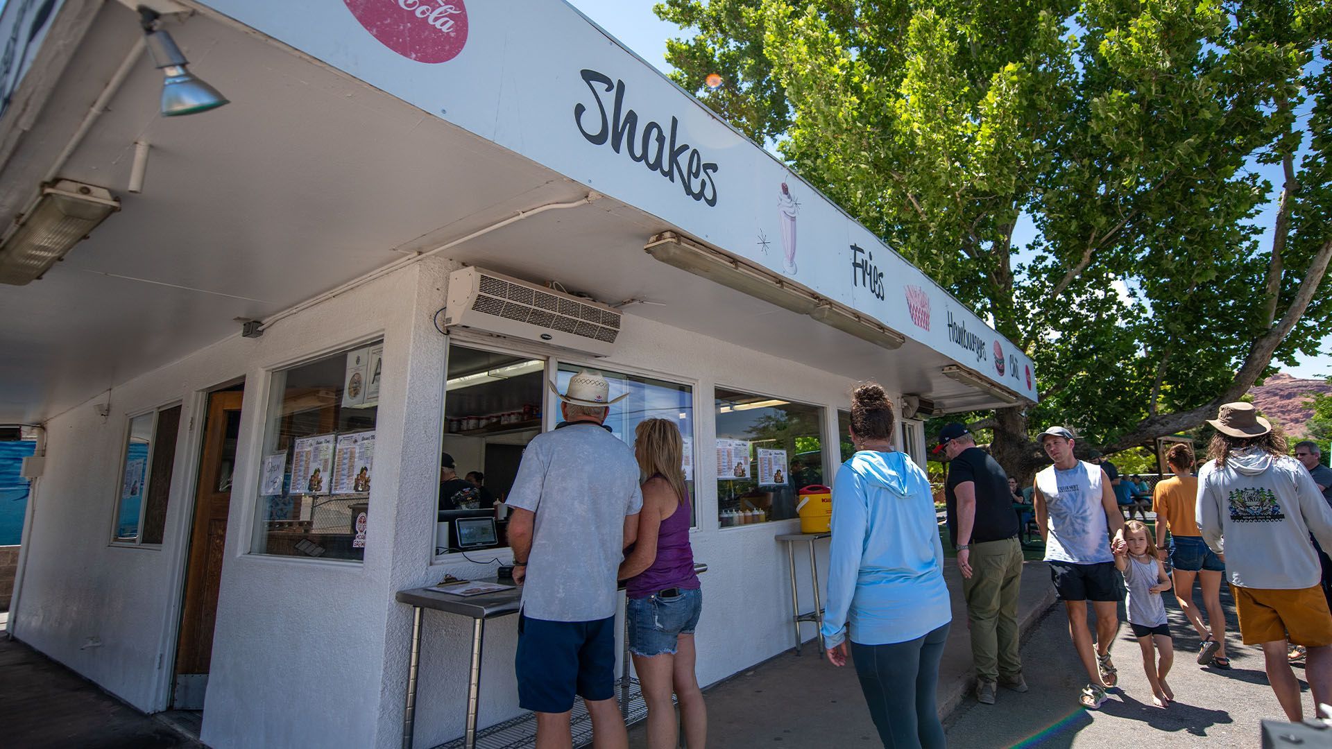 A small white ice cream shop with customers at a walk up window, sign says shakes, fries, burgers.
