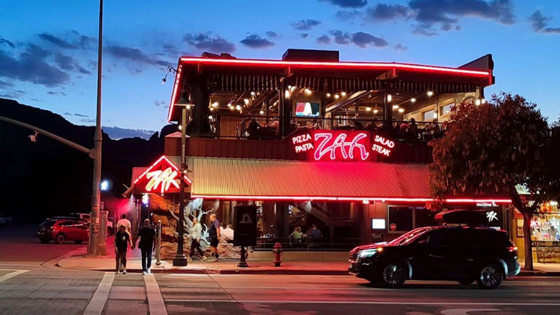 A two-story restaurant with neon lights, people dining, and cars on the street at dusk.