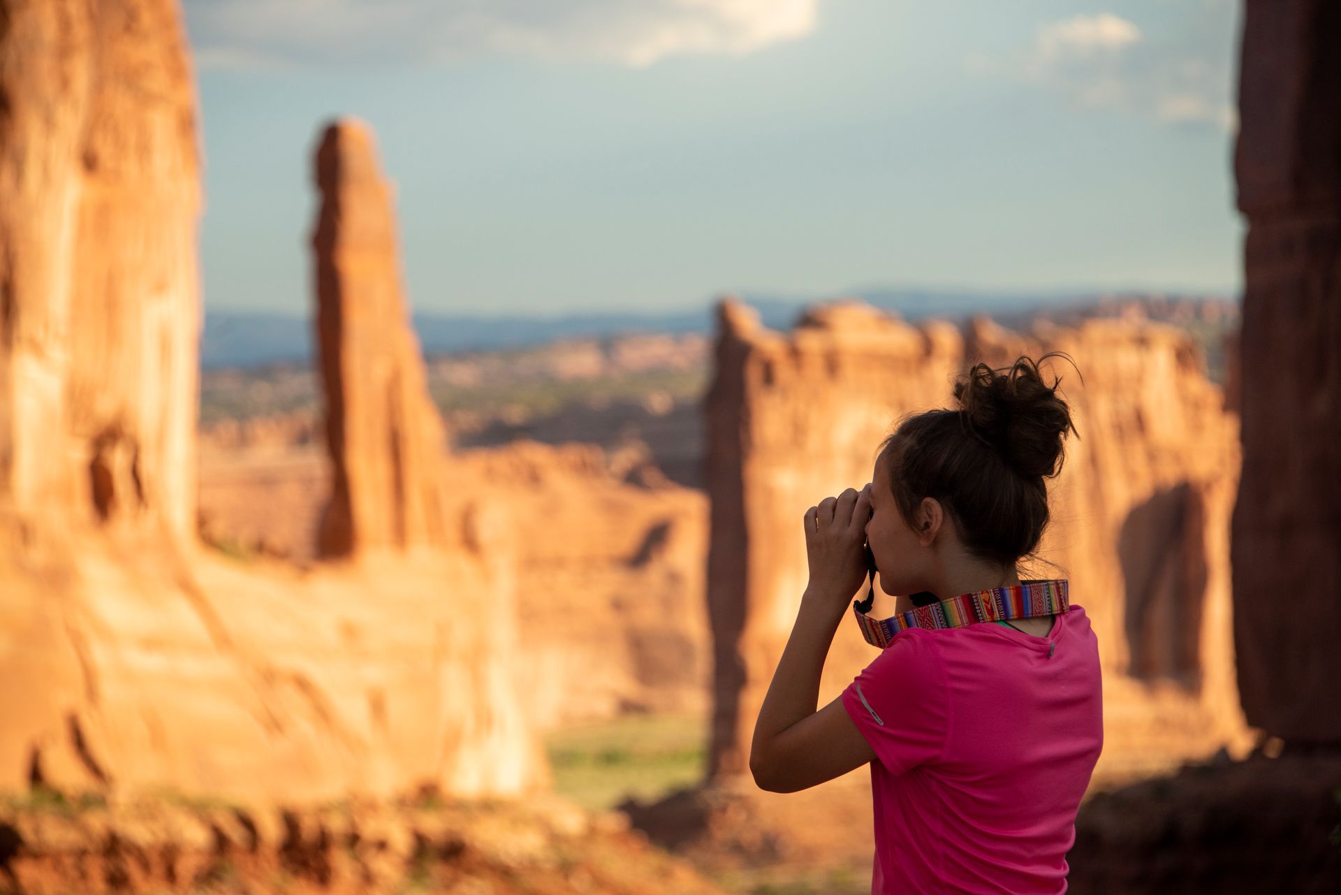 Woman looking through binoculars at rock formations. Outdoors, wearing a pink shirt.
