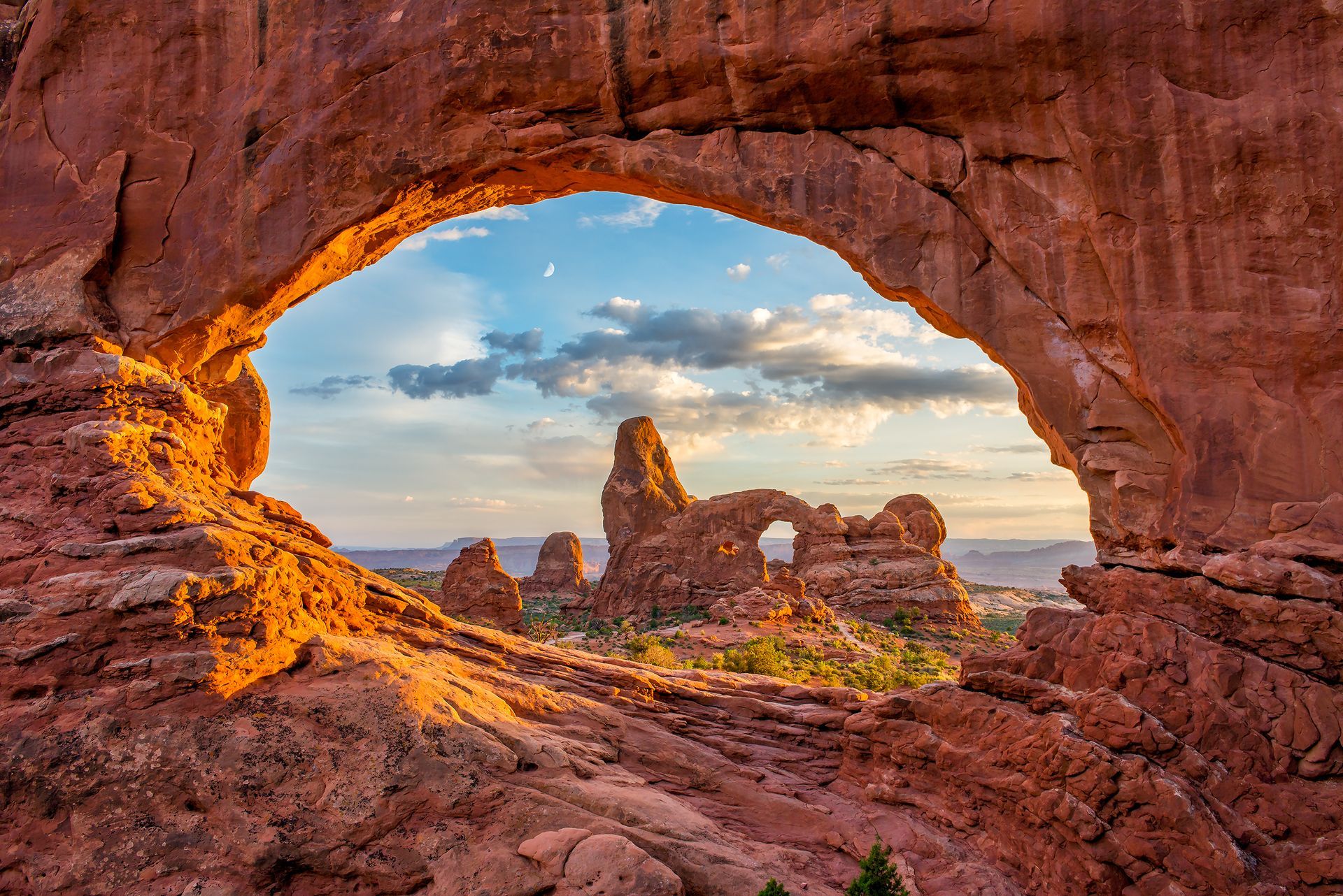 Red rock arch framing a view of desert landscape at sunset, including other rock formations.