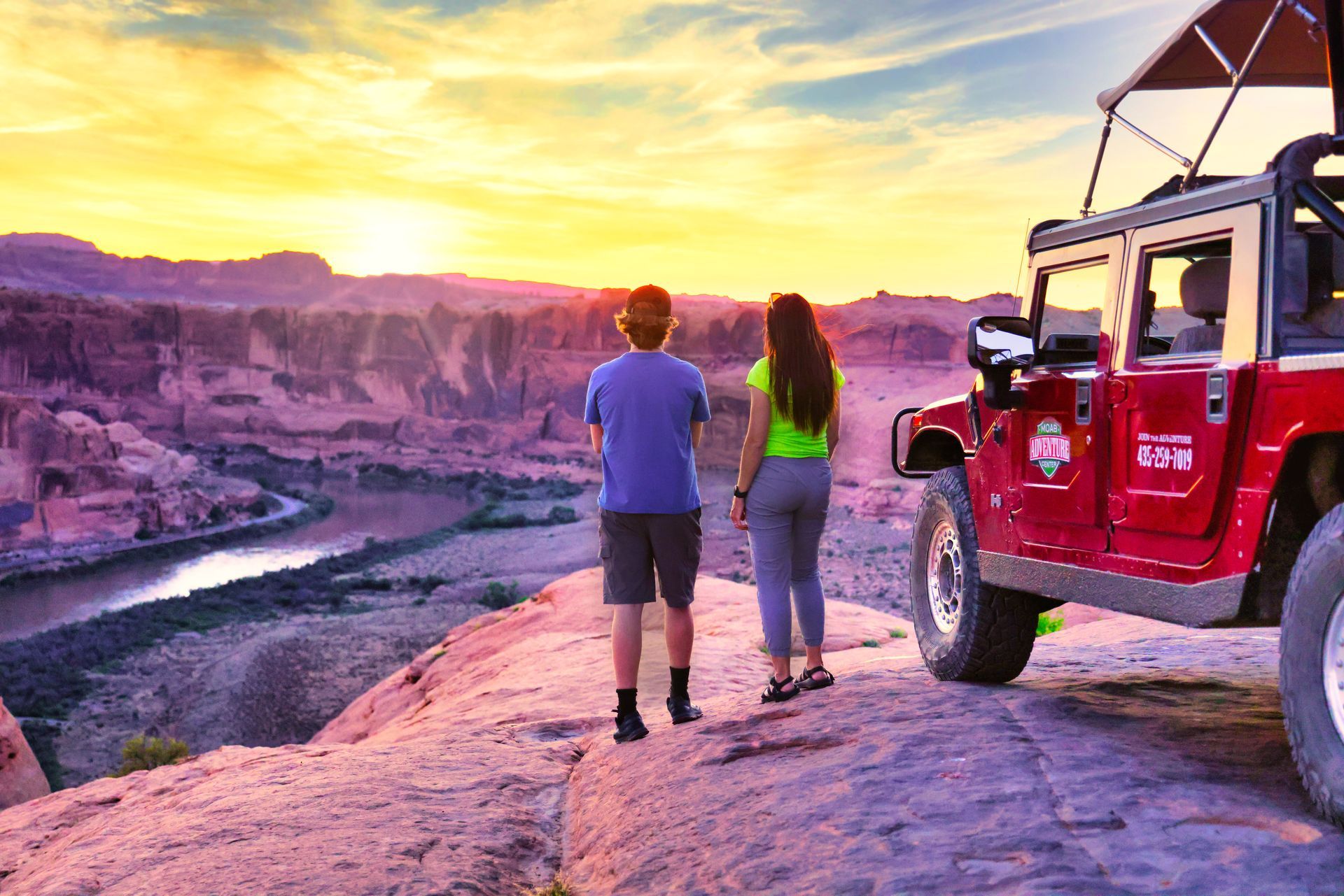 Two people watching a sunset over a canyon, next to a red Hummer.
