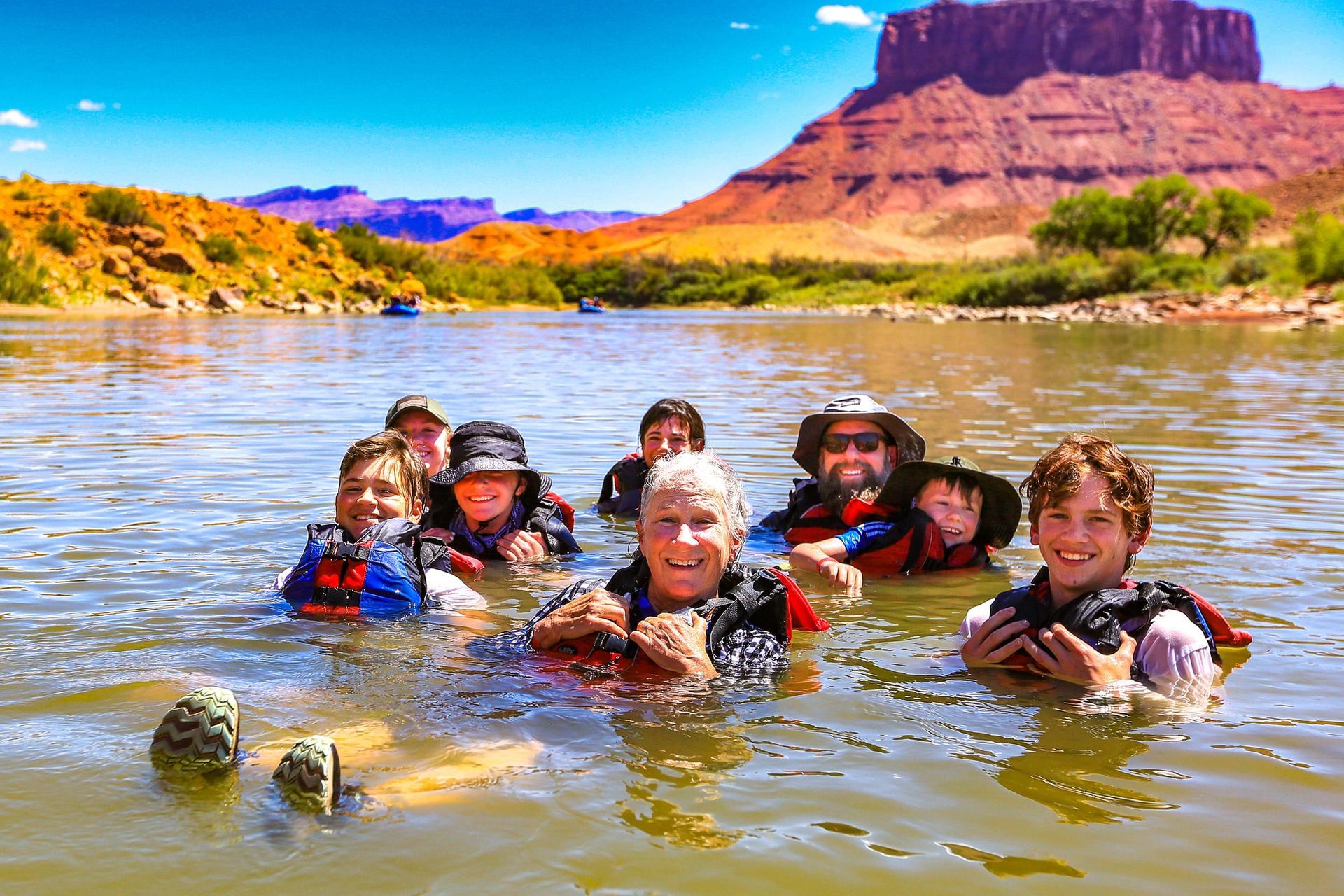 Group of people swimming in a river with a red rock mountain in the background on a sunny day.