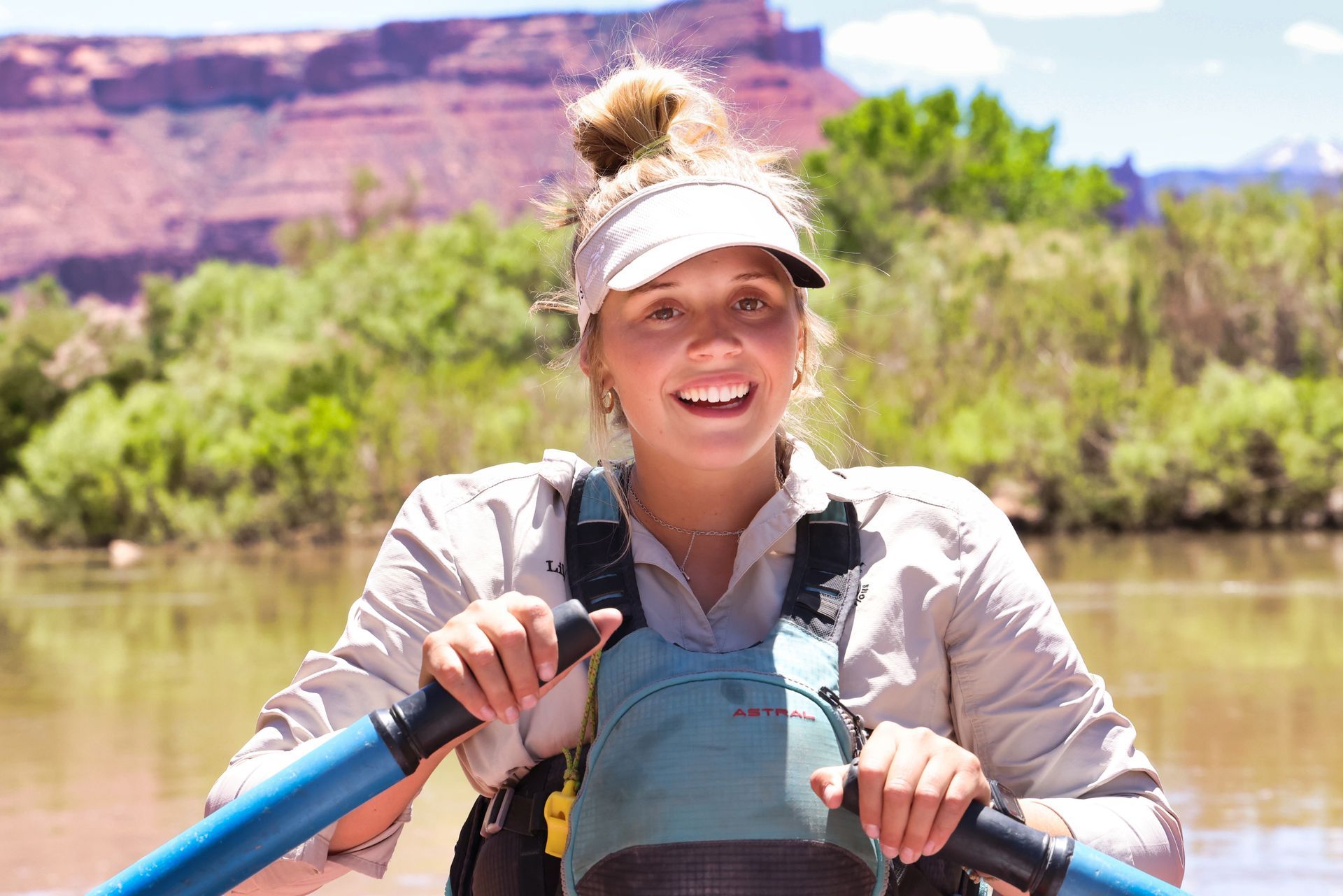 Female river guide smiles on a boat, holding oars. River, mountains, and green trees visible in background.
