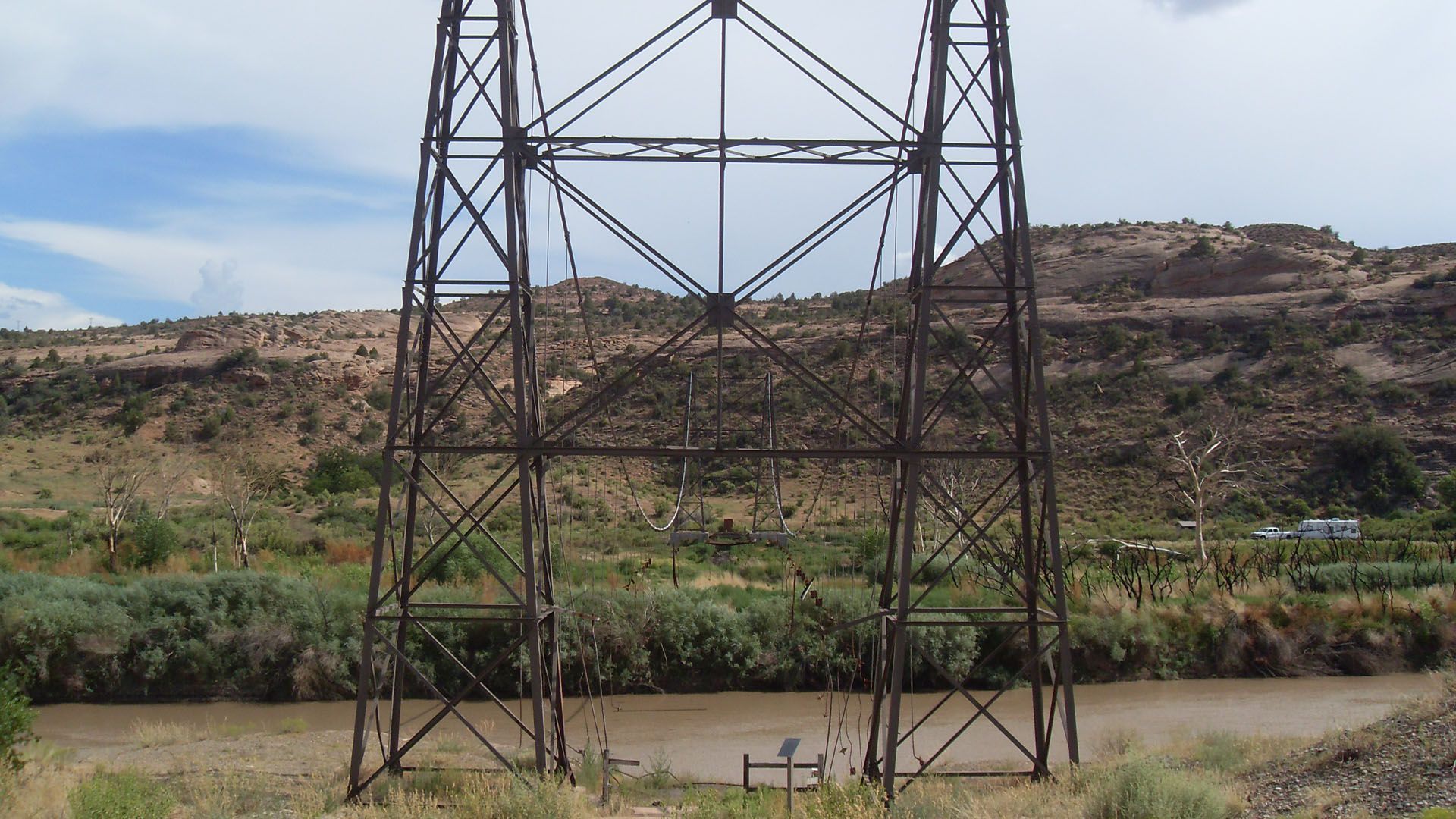 A large, metal power line tower stands over a muddy river, with a desert landscape in the background.