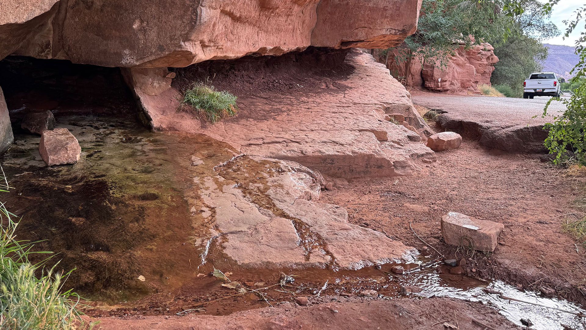 Water flows from a red rock overhang onto a cracked road, a truck is visible.