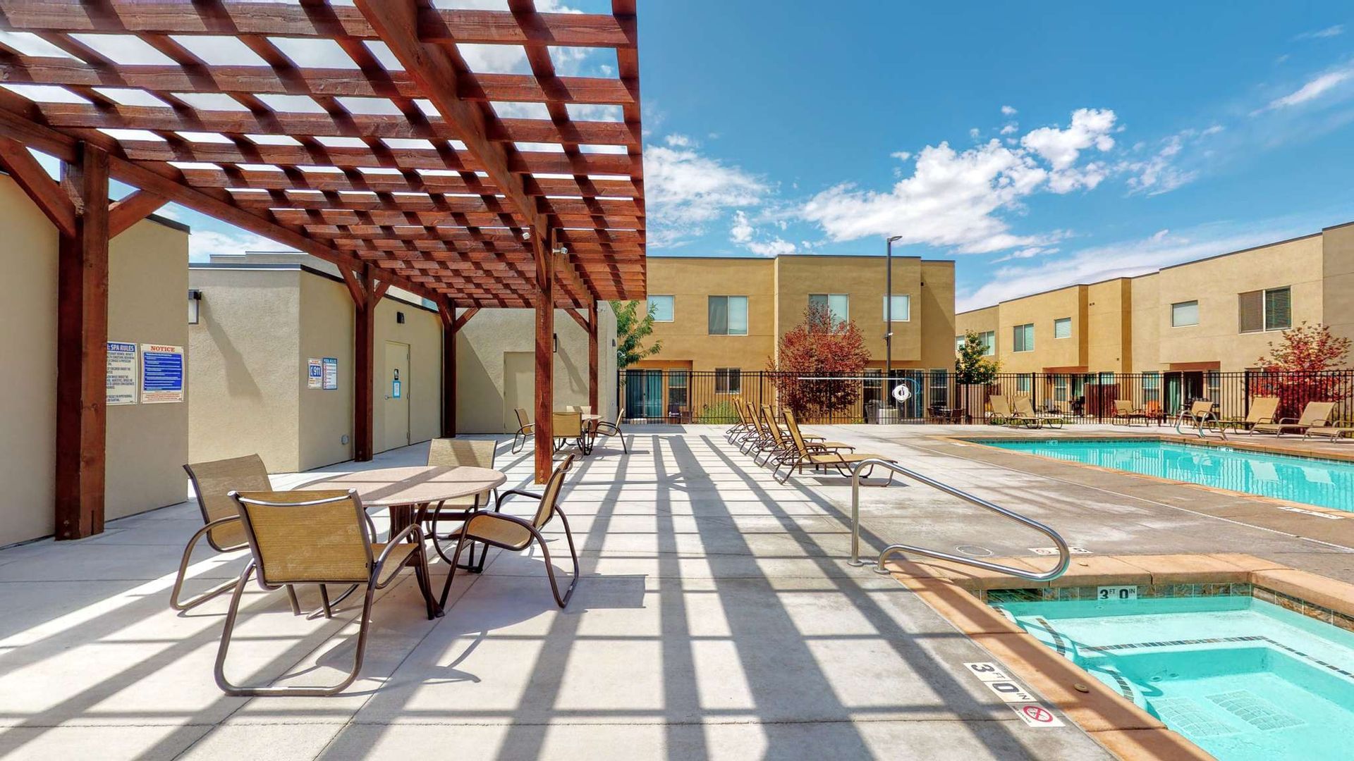 Outdoor pool area with pergola shade, tables, chairs, and a building in the background. Blue sky.
