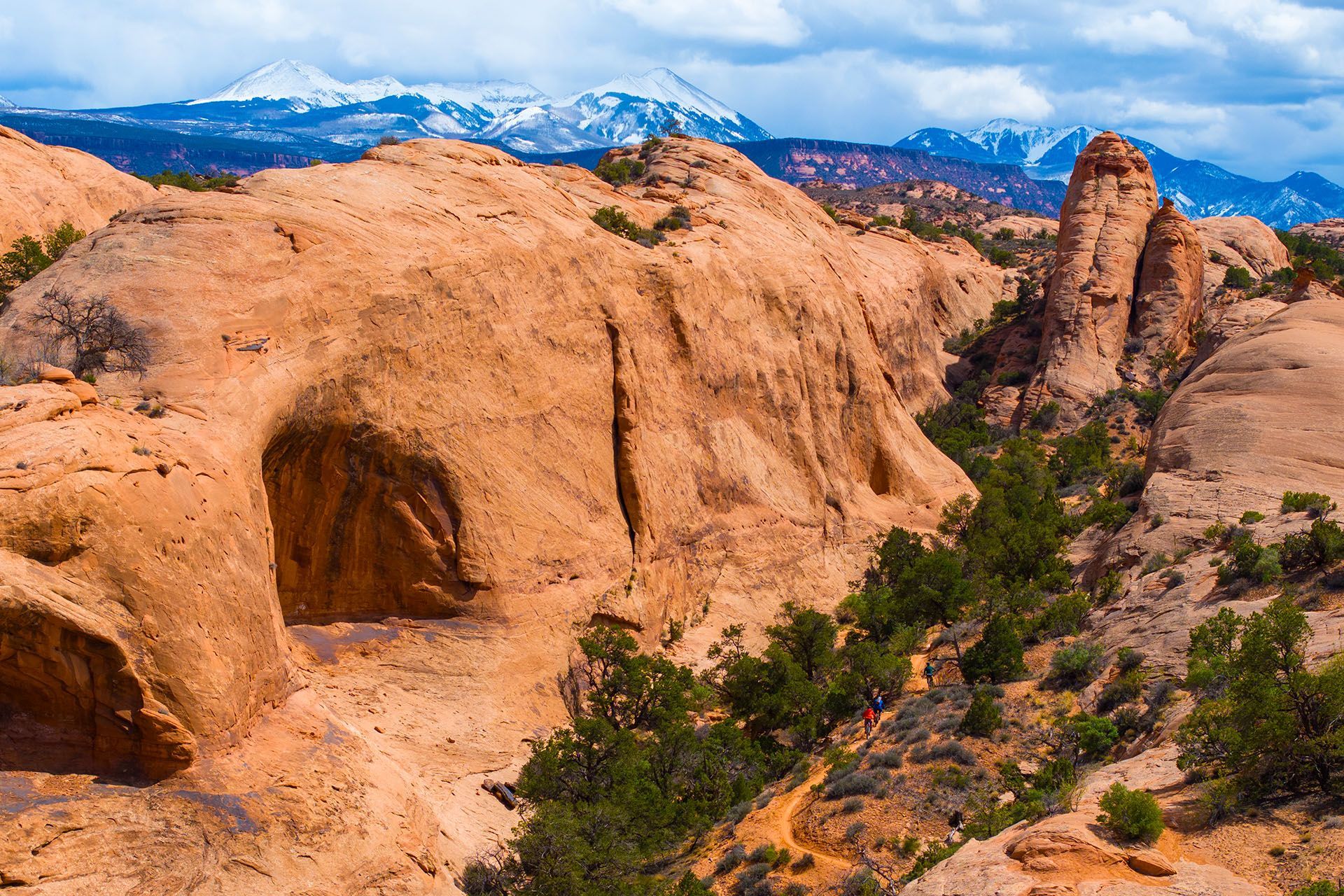 Red rock canyon landscape with arch, greenery, and snow-capped mountains in the distance.