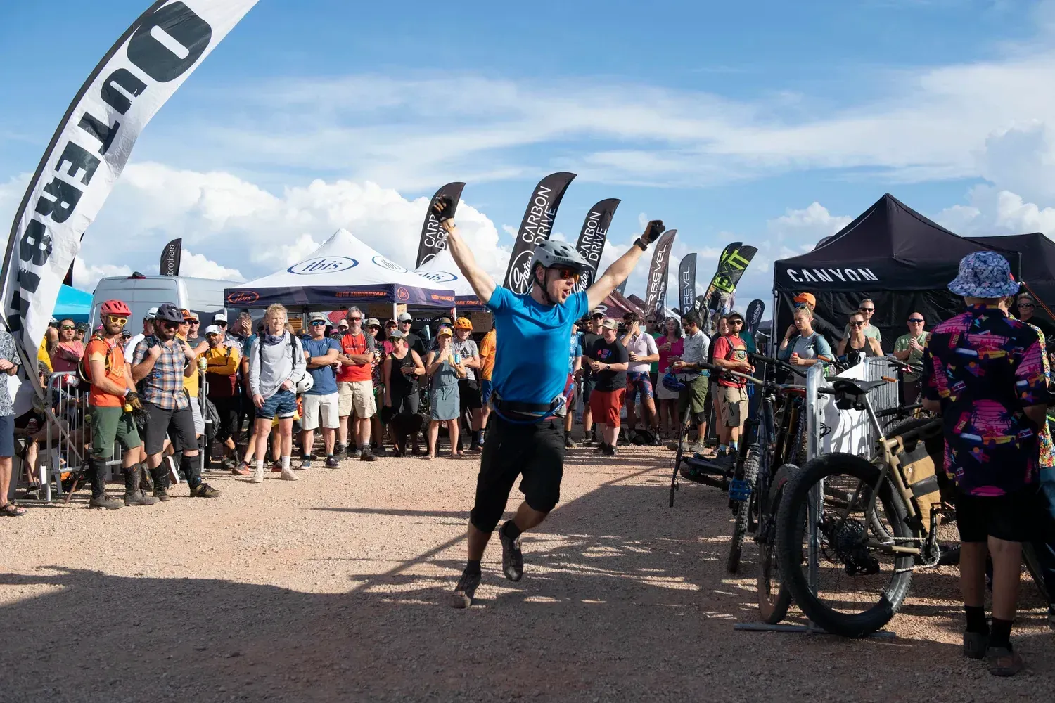 Man celebrates victory at a bike race finish line, arms raised. Crowd watches under sunny sky.