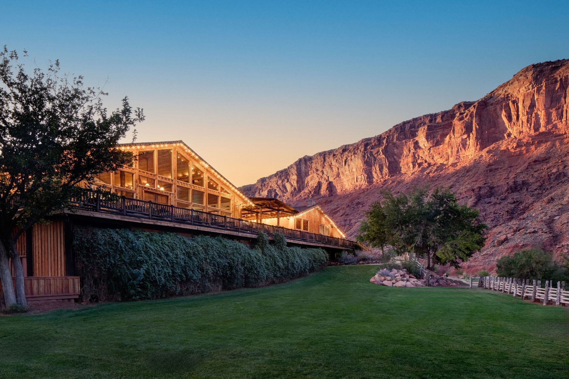 A lodge with a green lawn sits at the base of a reddish mountain under a blue sky at sunset.