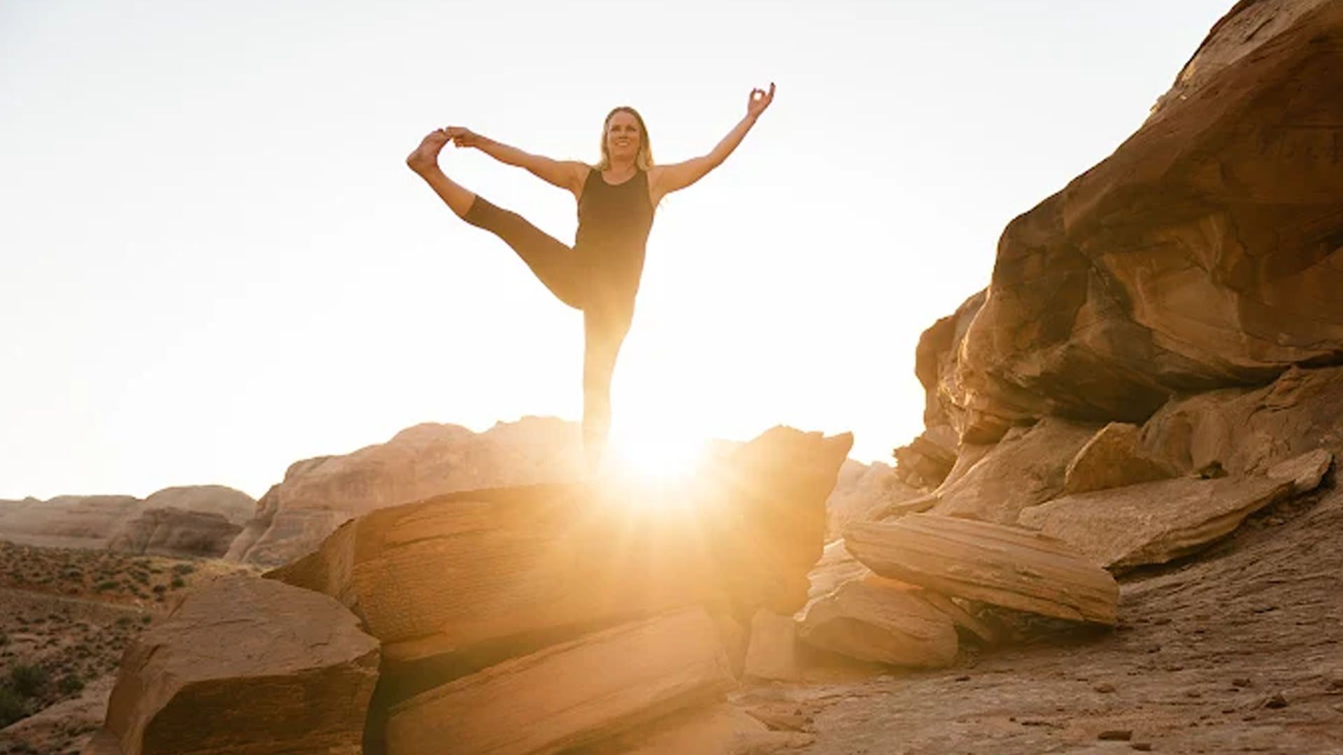 Woman performs a yoga pose on a large sandstone rock with sunrise behind her.